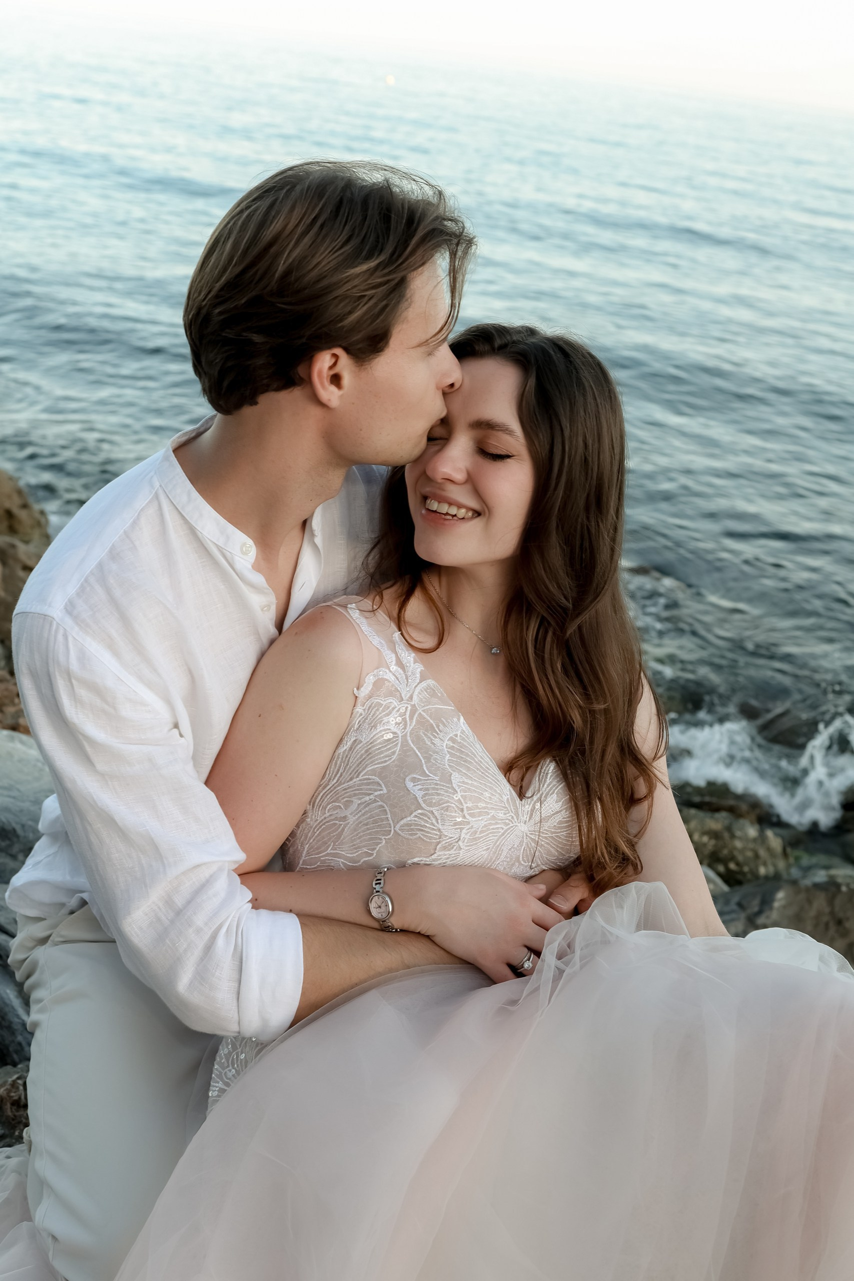 Bräutigam küsst zart die Schläfe der Braut, sie lächelt glücklich, Meer im Hintergrund, After Wedding Shooting Spanien