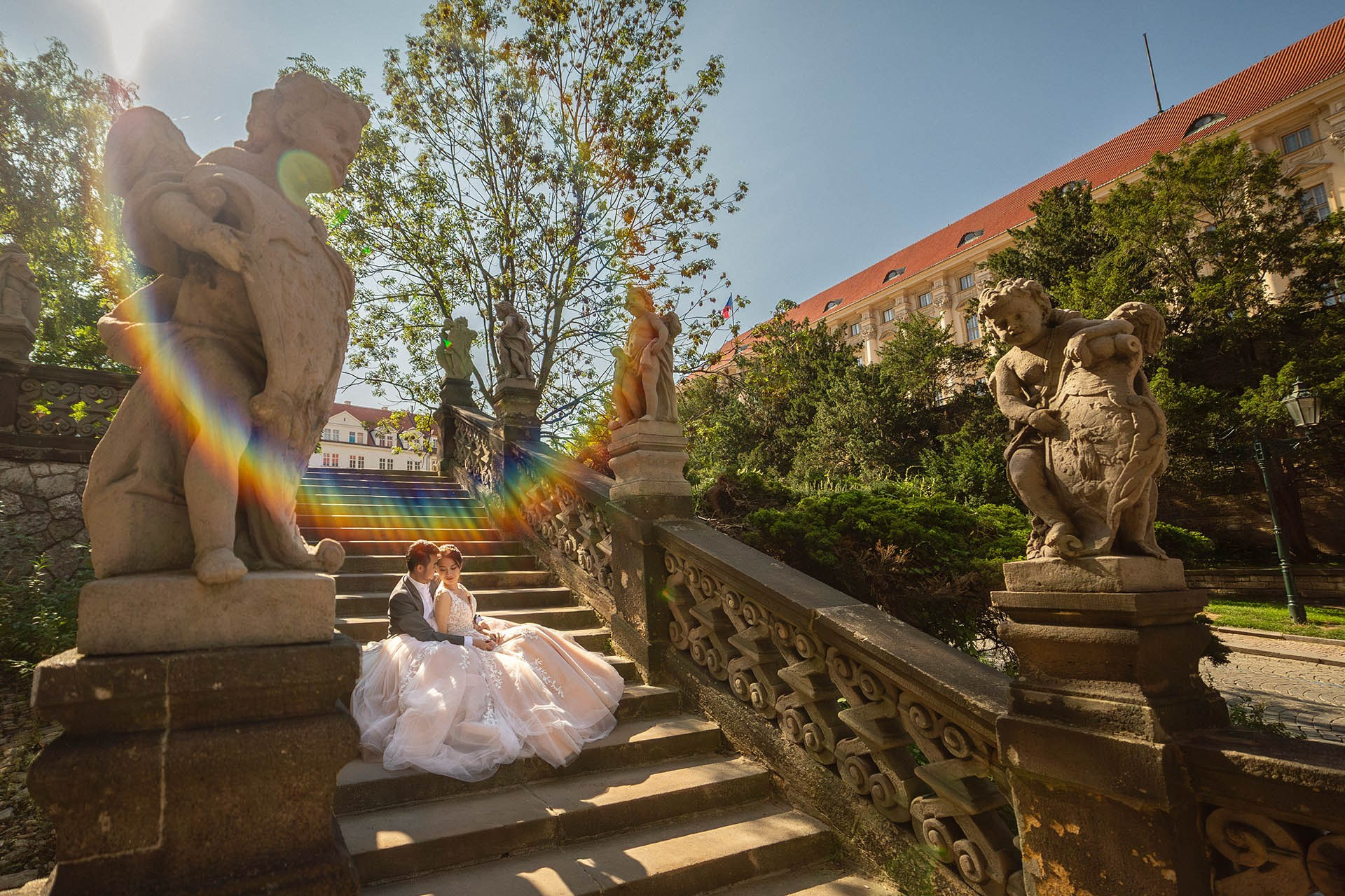 A couple sit among historic statues as the sun flares vividly above in the historic Loreto district of Prague Castle.