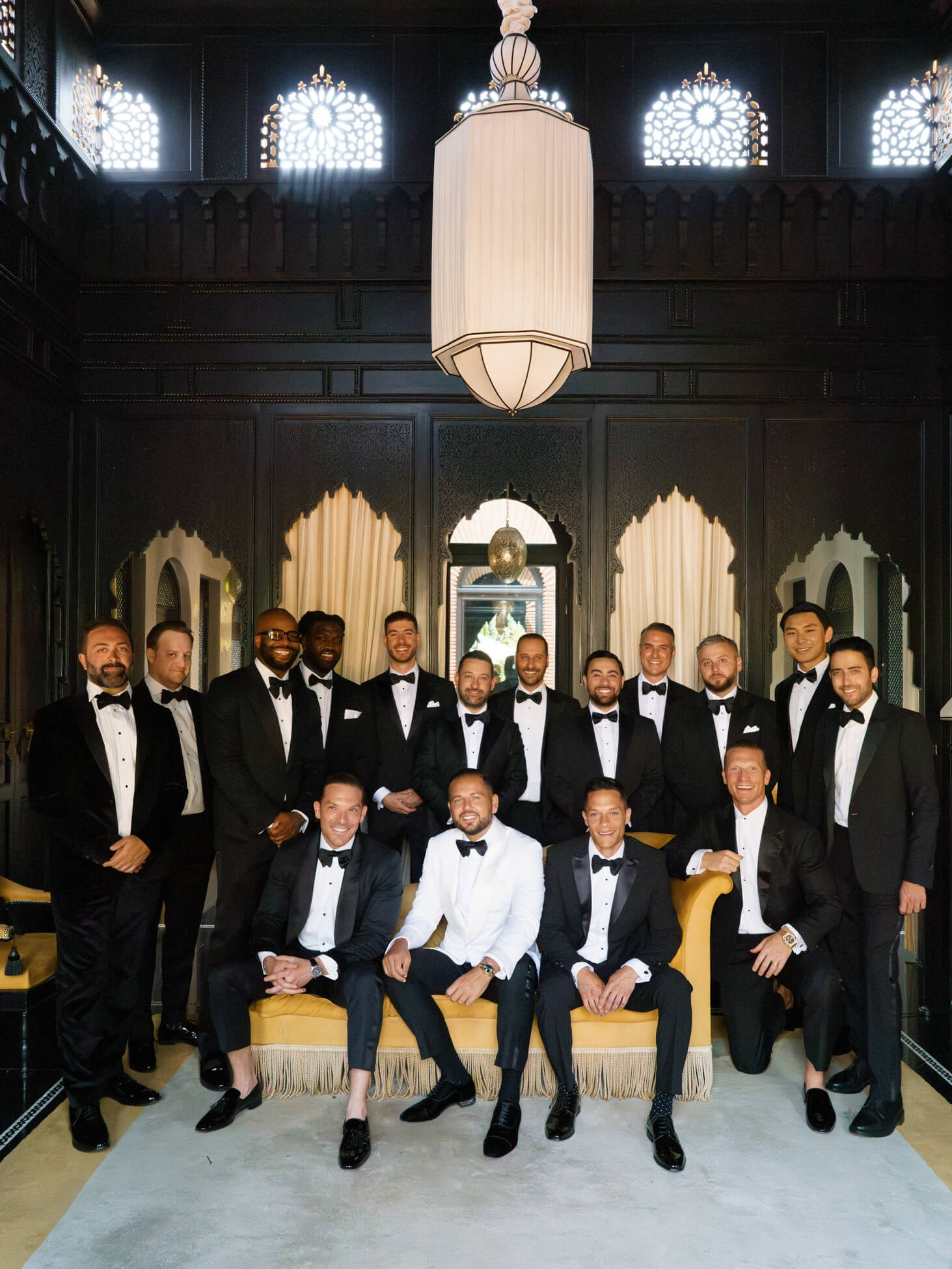 Groom in white tuxedo jacket and groomsmen in black tie posed inside an ornate Moroccan riad with carved arched windows and a pendant chandelier