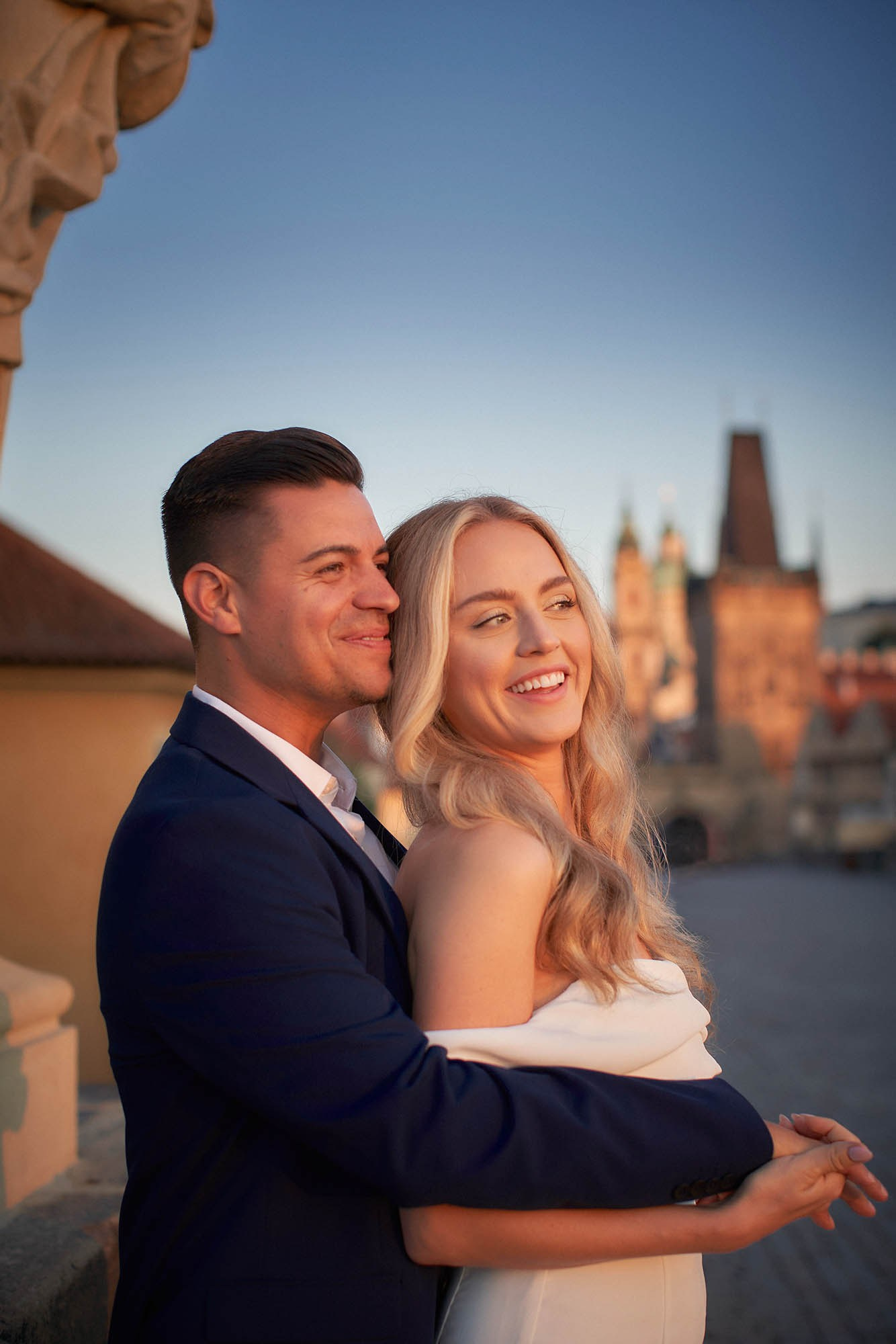 Joyful engaged couple faces glowing in early sunrise on Charles Bridge, Prague.