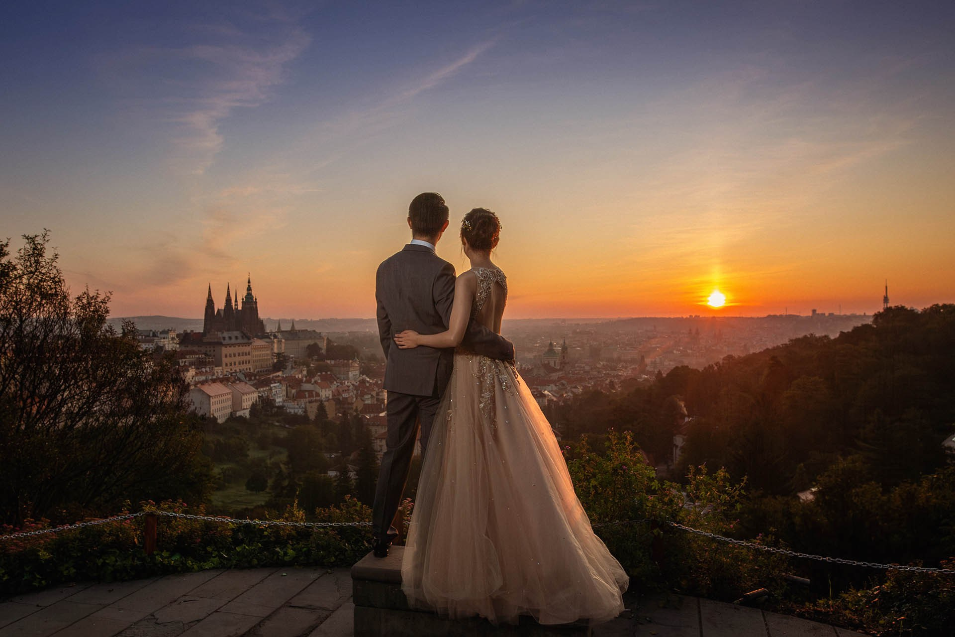 As the sun breaks above the skyline of Prague, a young couple are nearly silhouetted against the scenery below.