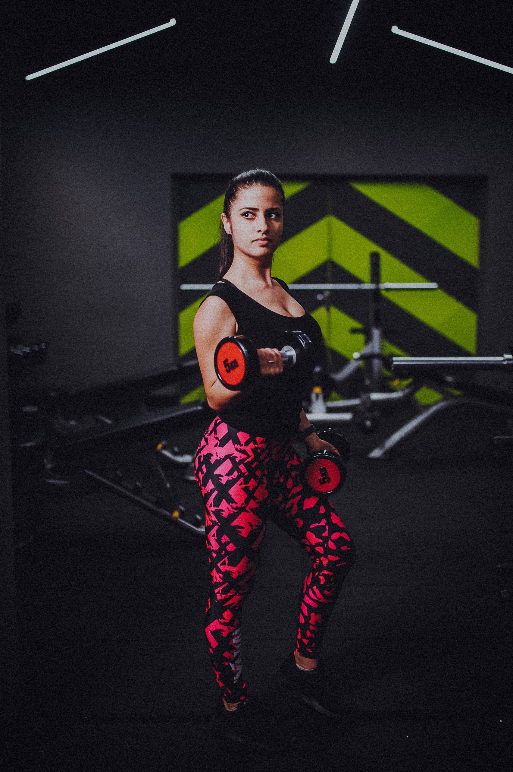 Antrenoare în maiou negru și colanți cu imprimeu roz, cu o ganteră ridicată și cealaltă lângă corp, în sala de fitness.