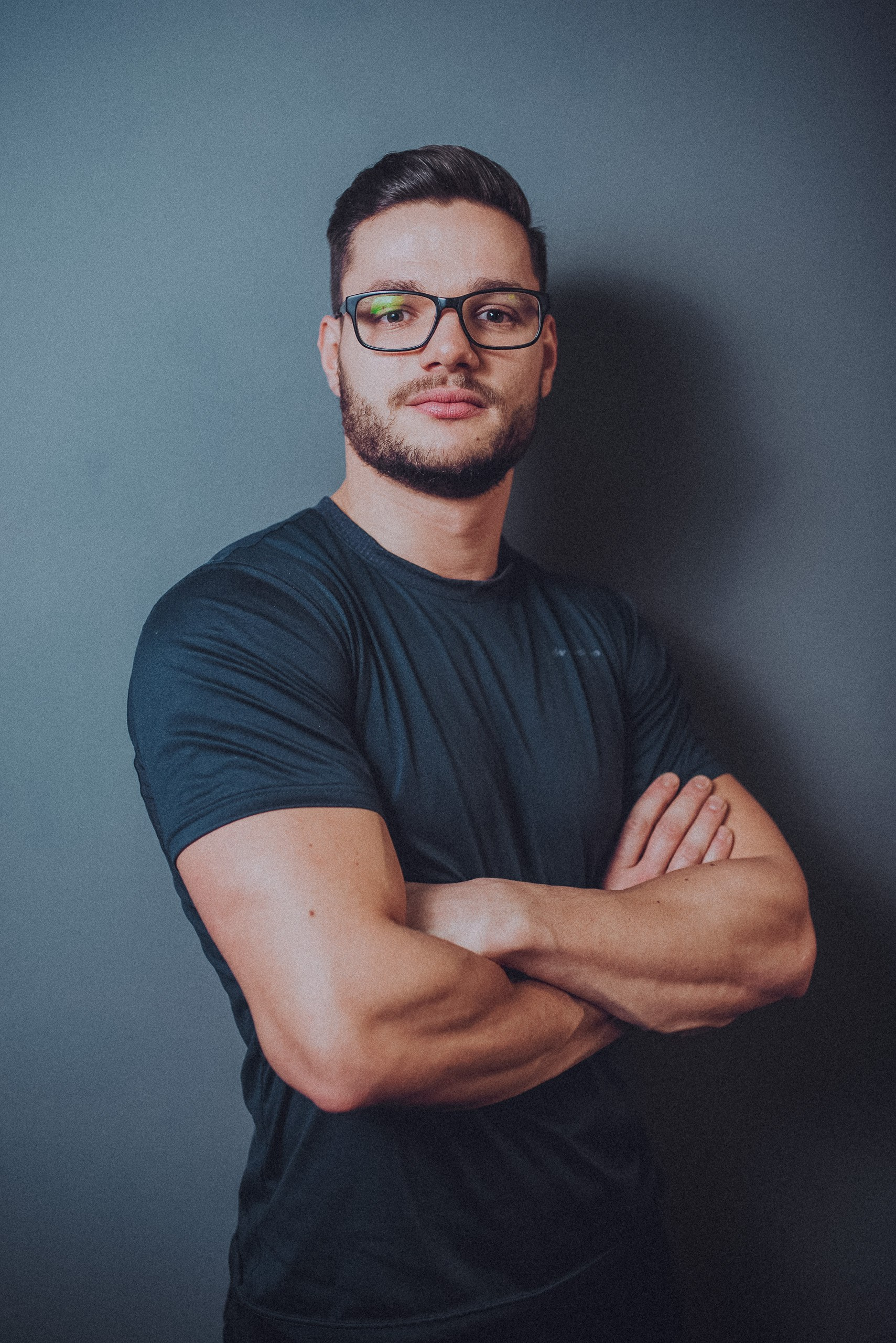 Antrenor cu ochelari și tricou închis la culoare, privind spre cameră cu brațele încrucișate, pe fundal gri.