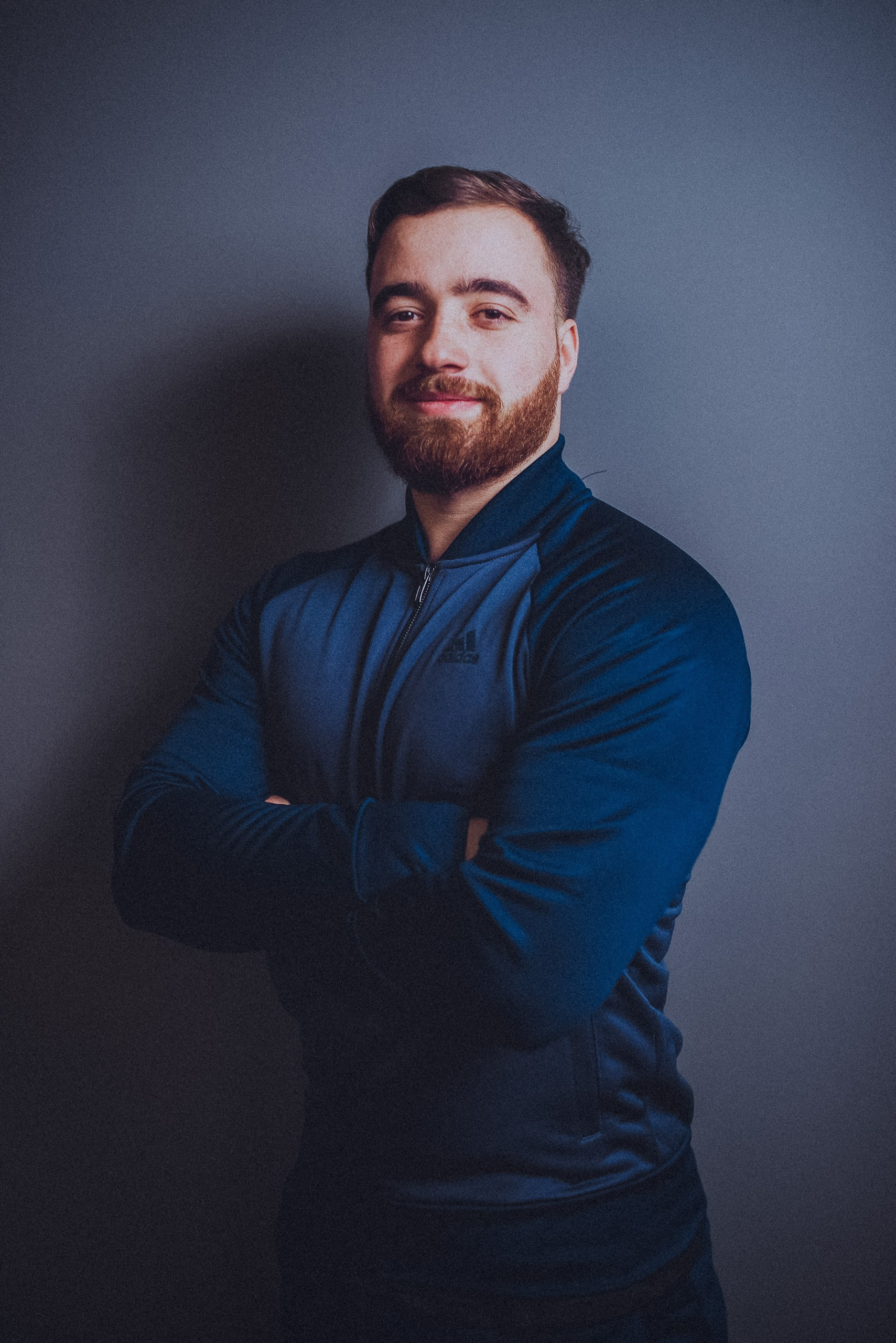 Antrenor cu barbă, în bluză sport albastră, cu brațele încrucișate și un zâmbet discret, pe fundal gri.