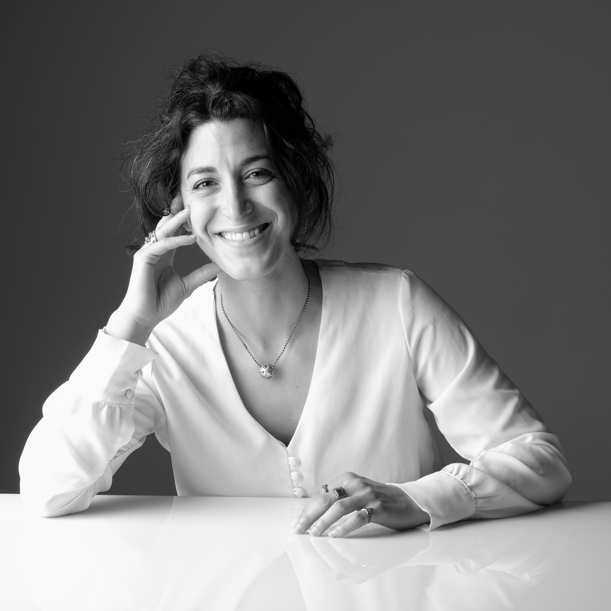 A black and white photo of Julie Nakzi sitting behind a table with one arm next to her head.
