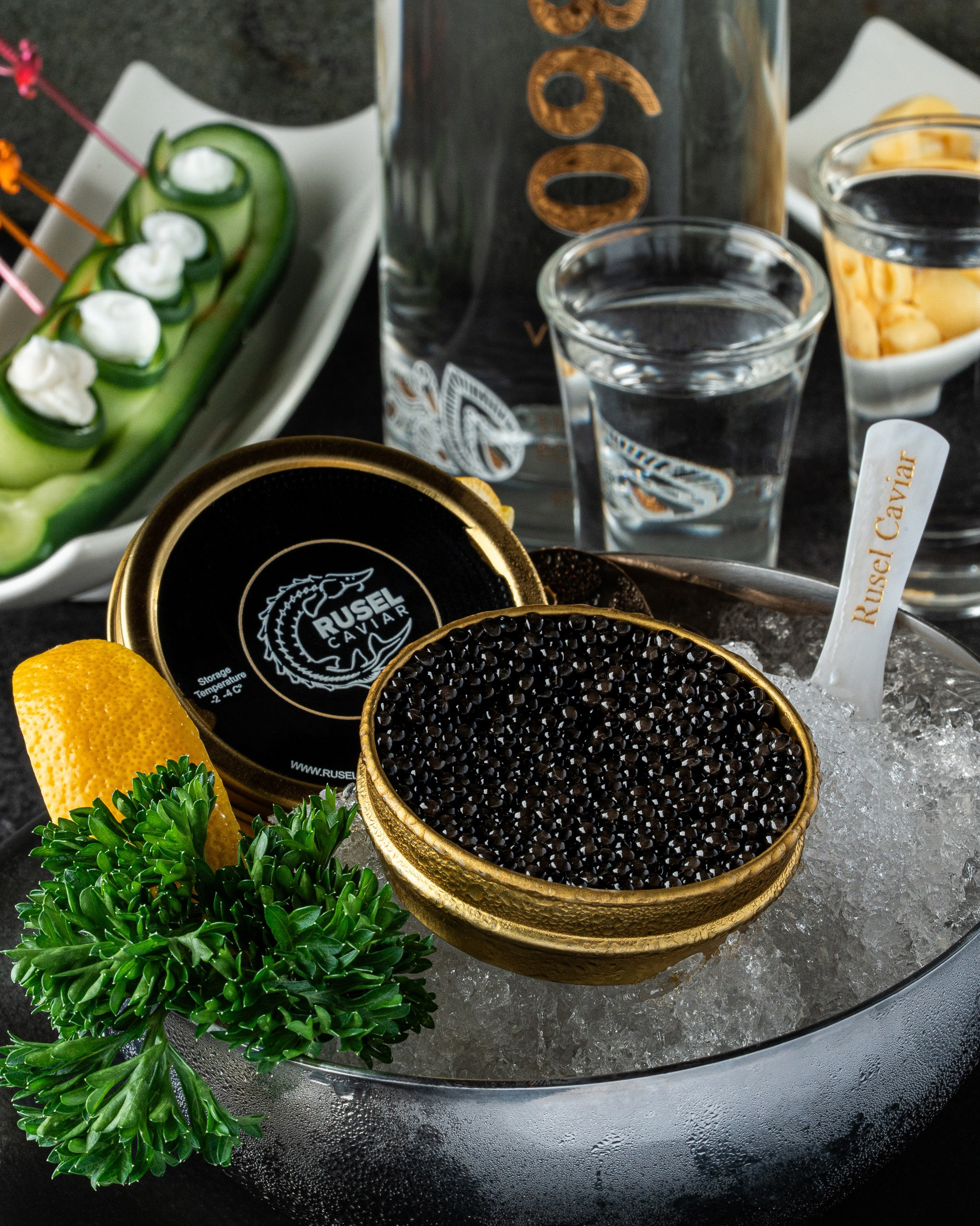 Rusel Caviar black caviar served on ice with a lemon slice and parsley; Xan 1860 vodka and appetizers in the background.