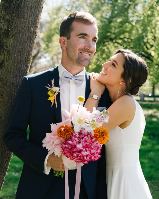Die Braut blickt auf ihren Bräutigam, während er Hochzeitsblumen unter dem Baum im Englishen Garten hält München