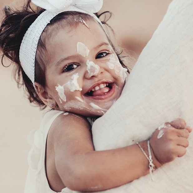 Birthday girl with a white bow and cake cream on her face, smiling joyfully while hugging an adult during a beach birthday photoshoot in Puglia.