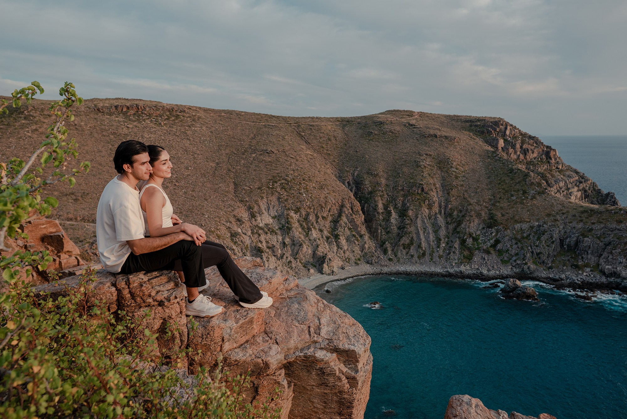 Couple sitting on a cliff in neutral-toned boots, looking at the ocean in Los Cabos