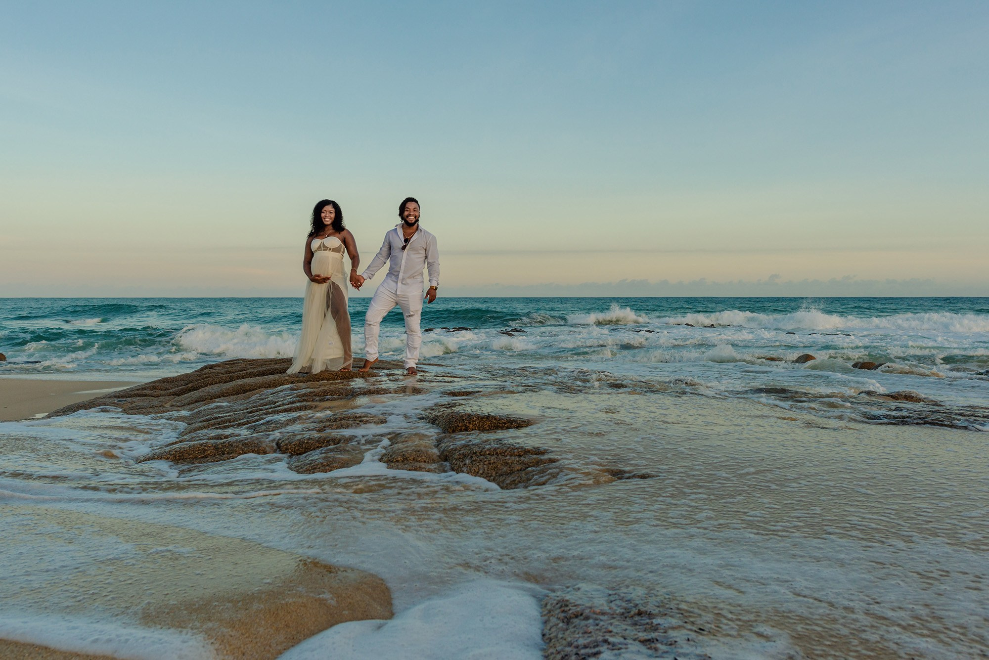 Pregnant couple in a white chiffon dress moving with the wind on rocks by the beach