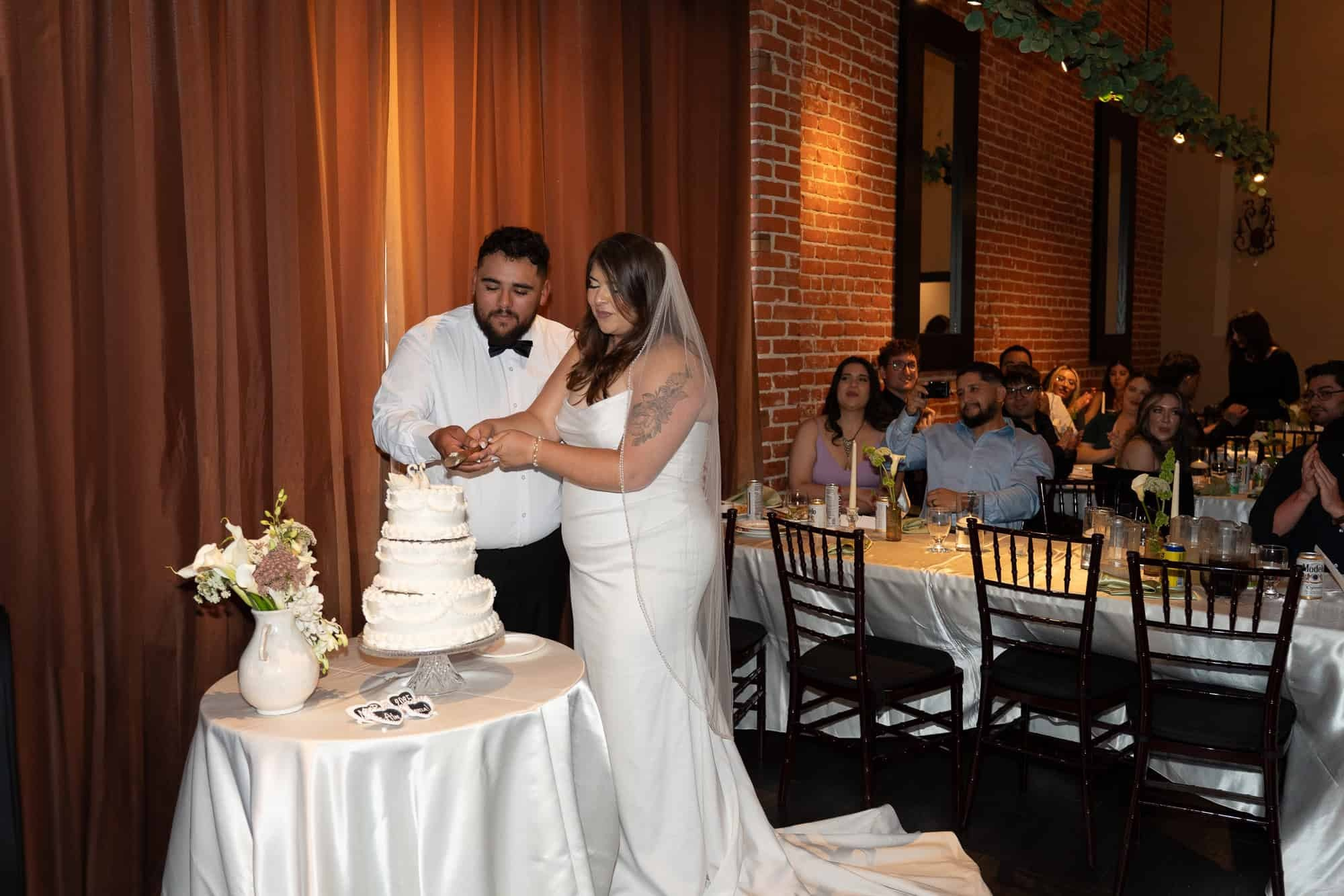 Bride and groom cutting their wedding cake at reception, surrounded by family and friends inside cozy restaurant in Pasadena with brick walls