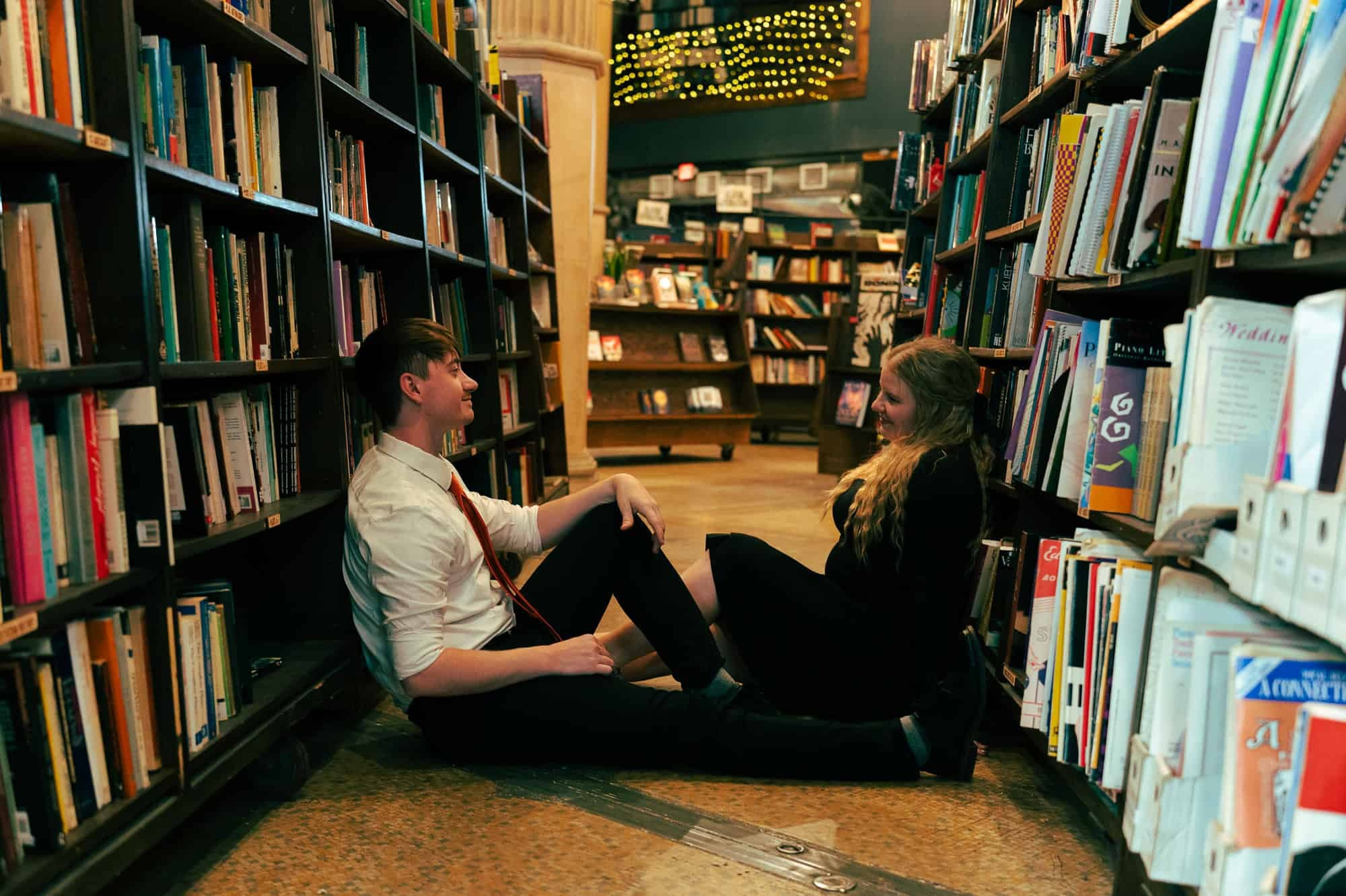 Engaged couple relaxing on the floor between tall bookshelves at The Last Bookstore in Downtown Los Angeles, sharing smiles and quiet connection during a cinematic engagement session with cozy, literary vibes