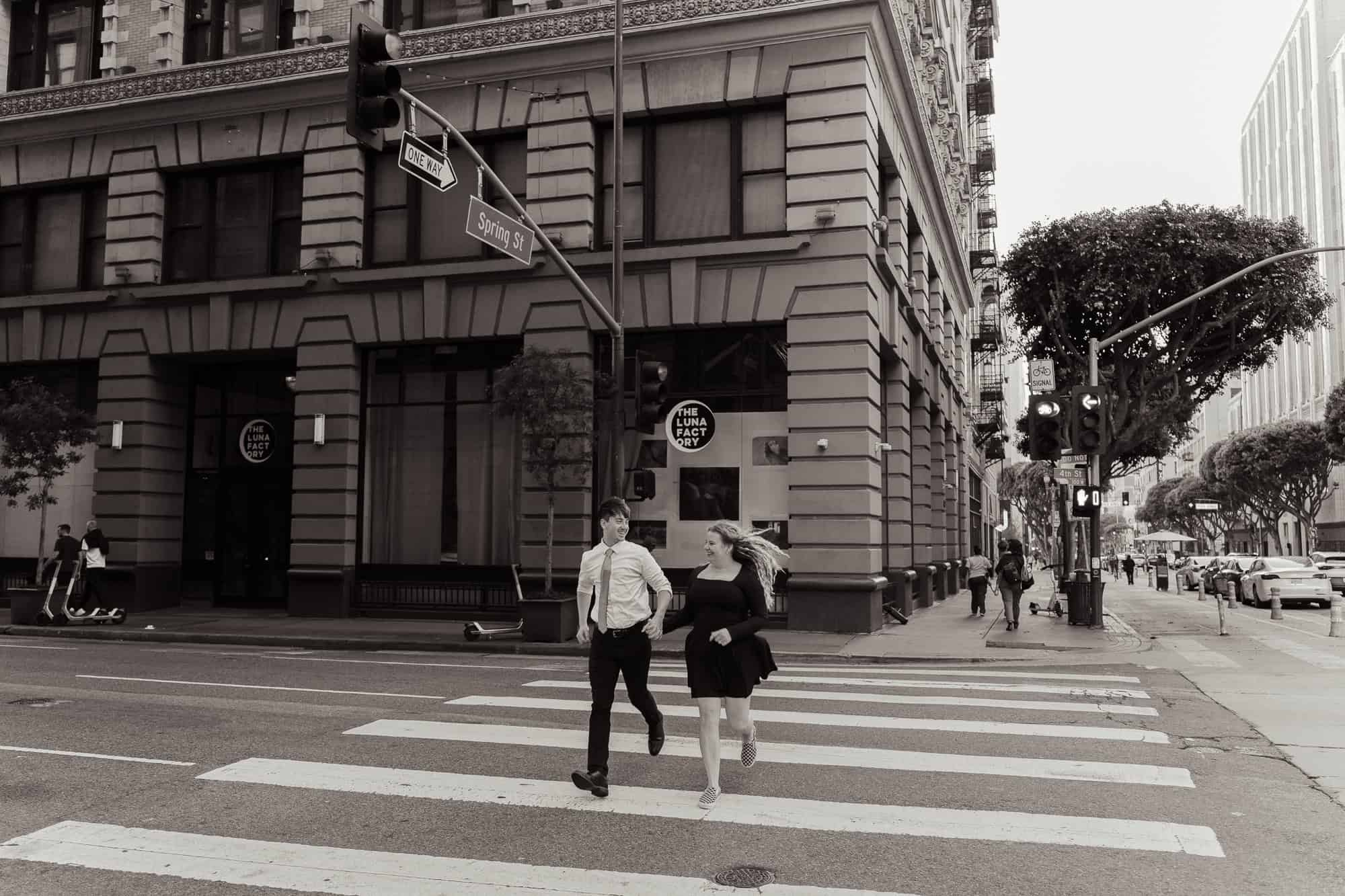 Candid black and white photo of a couple joyfully running through the streets of Downtown Los Angeles, surrounded by historic architecture and the city’s vibrant energy