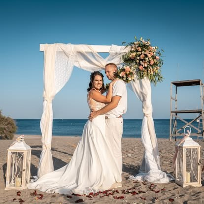 Romantische Strandhochzeit Foto: Brautpaar umarmt sich unter floral dekoriertem Hochzeitsbogen am Meer. Braut in weißem Spitzenkleid, Bräutigam in Strandkleidung. Dekoration mit Blumen, Stoff, Laternen und Blütenblättern auf Sand. Klare Himmel, Strand-Location, Hochzeitszeremonie am Meer.