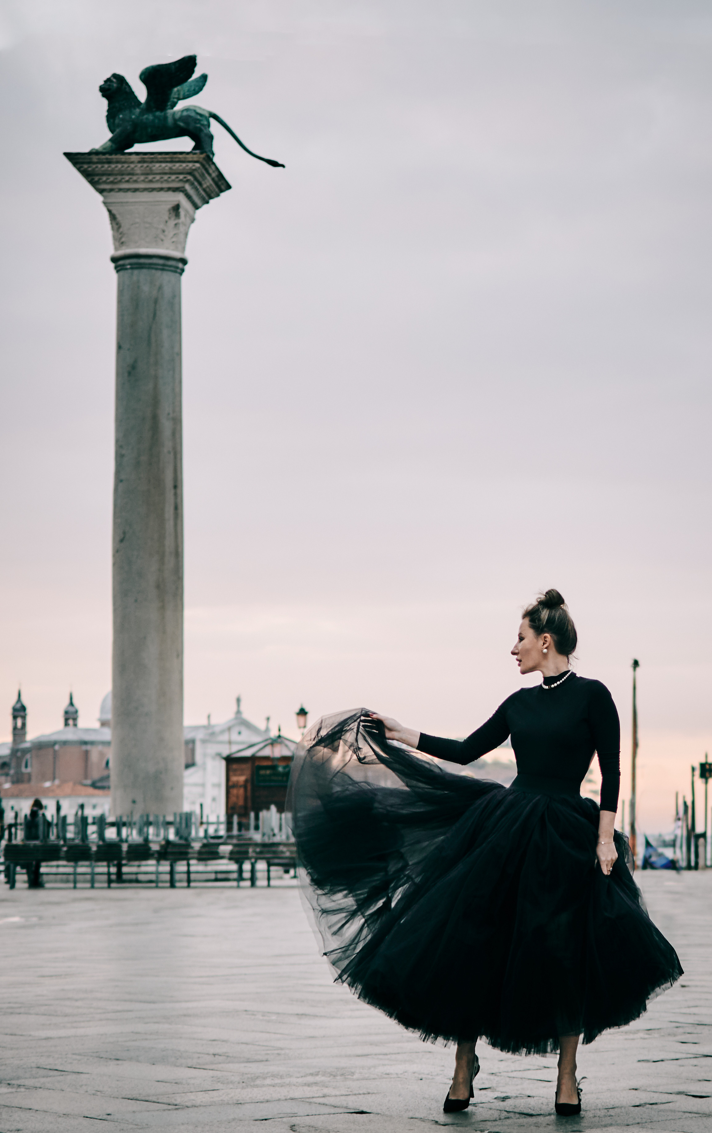 A young woman stands tall in front of the grand Columns of San Marco and San Todaro on the iconic San Marco Square in Venice. The woman's long hair flows freely in the gentle breeze as she gazes gracefully into the distance. The towering columns, adorned with intricate carvings, frame the woman beautifully, creating a striking contrast between the ancient architecture and the woman's youthful appearance. The warm, golden light of the setting sun bathes the scene, casting a soft glow on the woman's face and the majestic columns behind her. Fashion blogger shoot Venice. Portrait and Fashion Photographer in Venice, Italy. Instagram Photos with lifestyle photographer