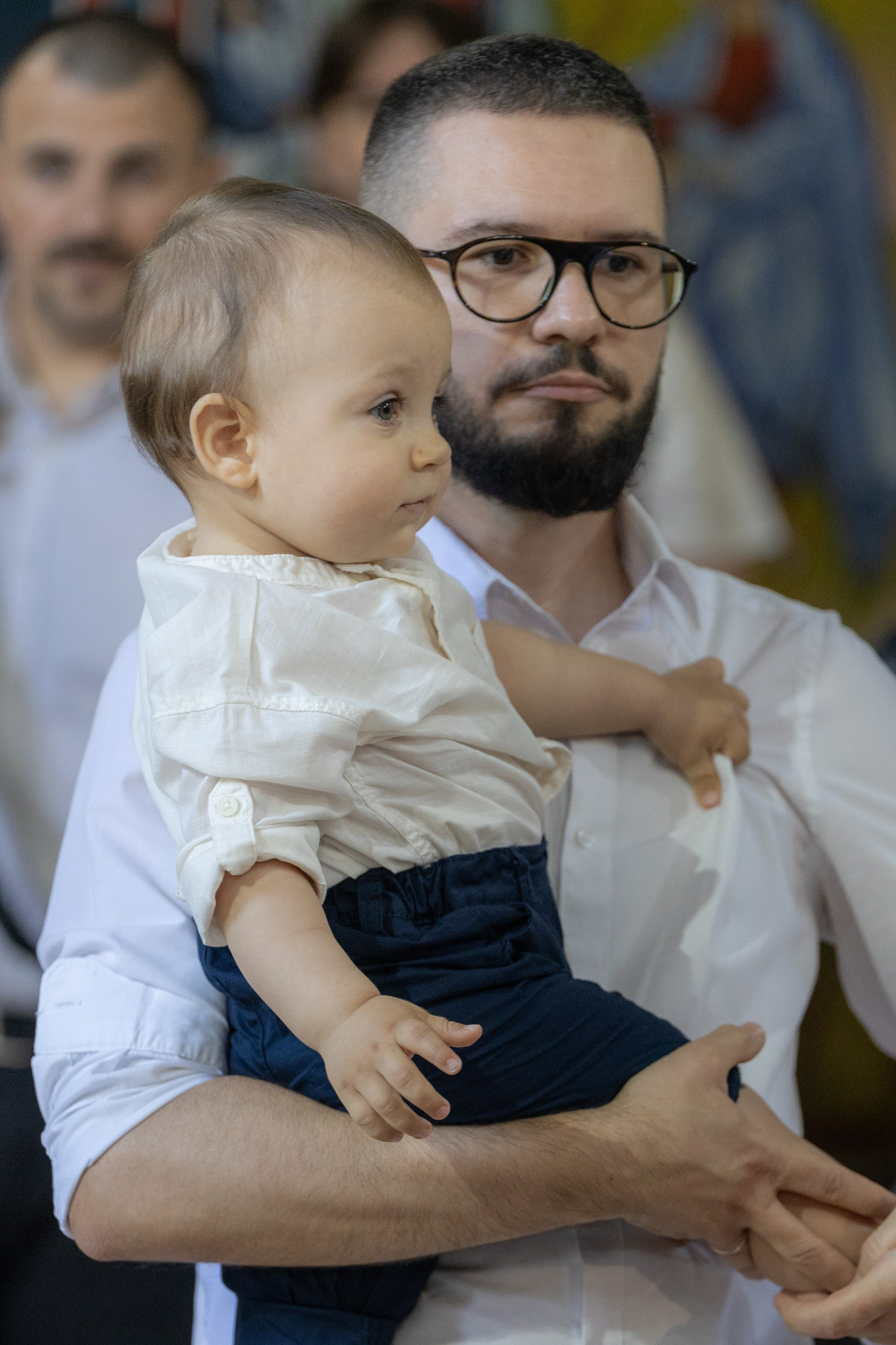 Svečan trenutak krštenja – kum s ljubavlju drži bebu dok sveštenik vrši obred. Srpska pravoslavna crkva Vavedenja Bogorodice u Zrenjaninu. 