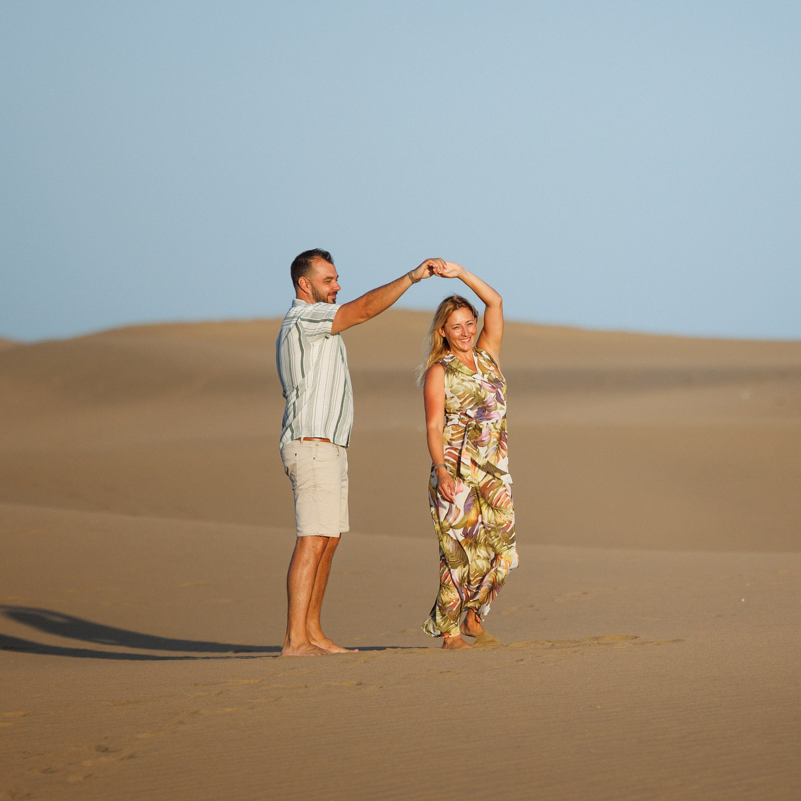 A man and woman dance barefoot on sand dunes under a clear blue sky, with the woman wearing a floral dress and the man in a short-sleeved shirt and shorts in Photoshoot Maspalomas