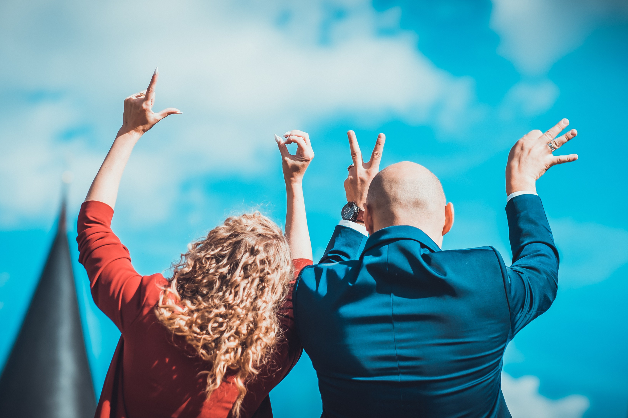 Zwei Personen, eine mit lockigem Haar in roter Kleidung und eine glatzköpfige Person im blauen Anzug, stehen Rücken zur Kamera und heben ihre Hände in die Luft, formen dabei Gesten. Der Himmel ist klar und blau, im Hintergrund sind die Spitzen von Türmen zu sehen