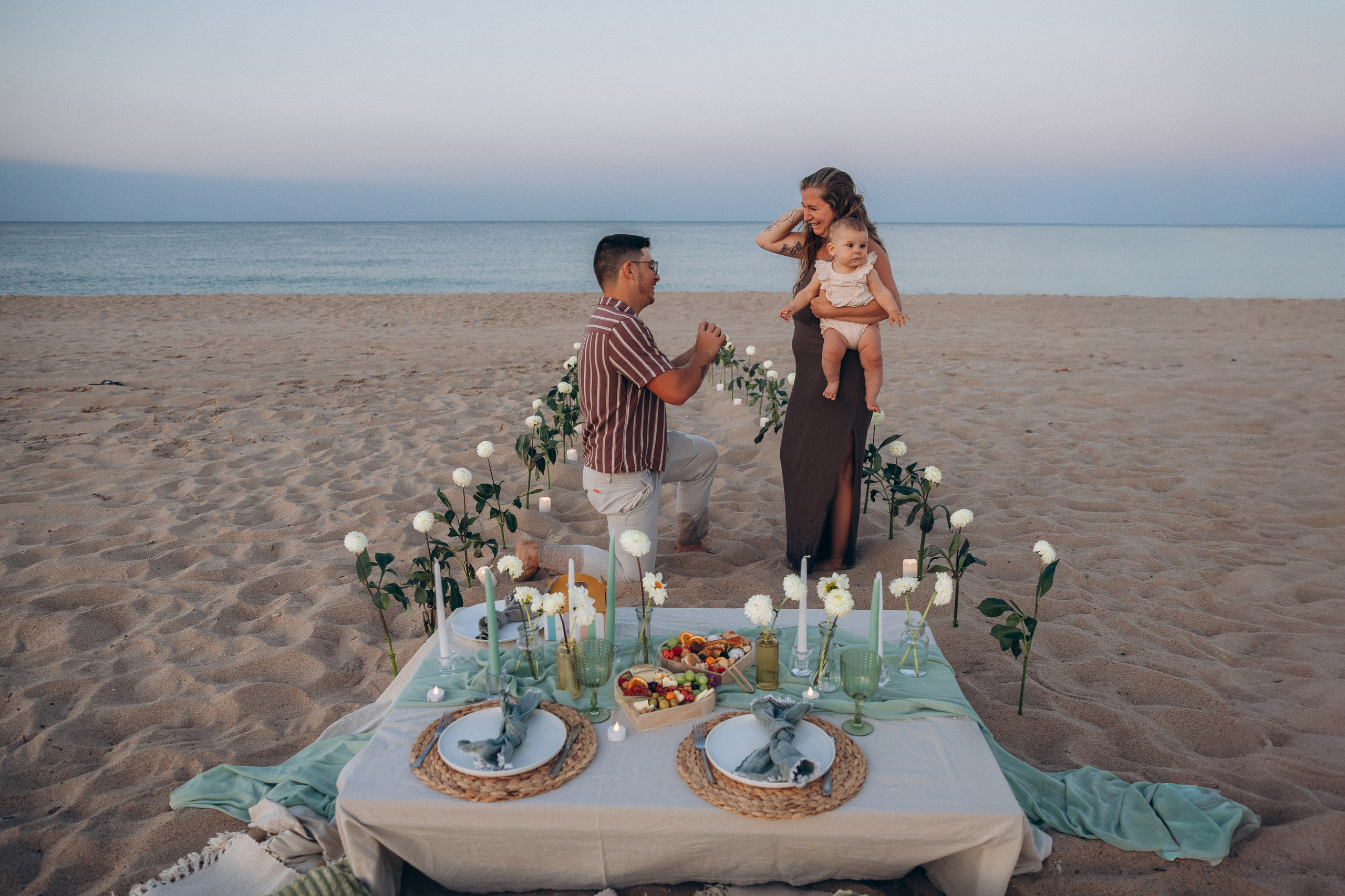 Surprise beach proposal in Valencia, with a man kneeling beside a flower-decorated picnic table as his partner reacts.
