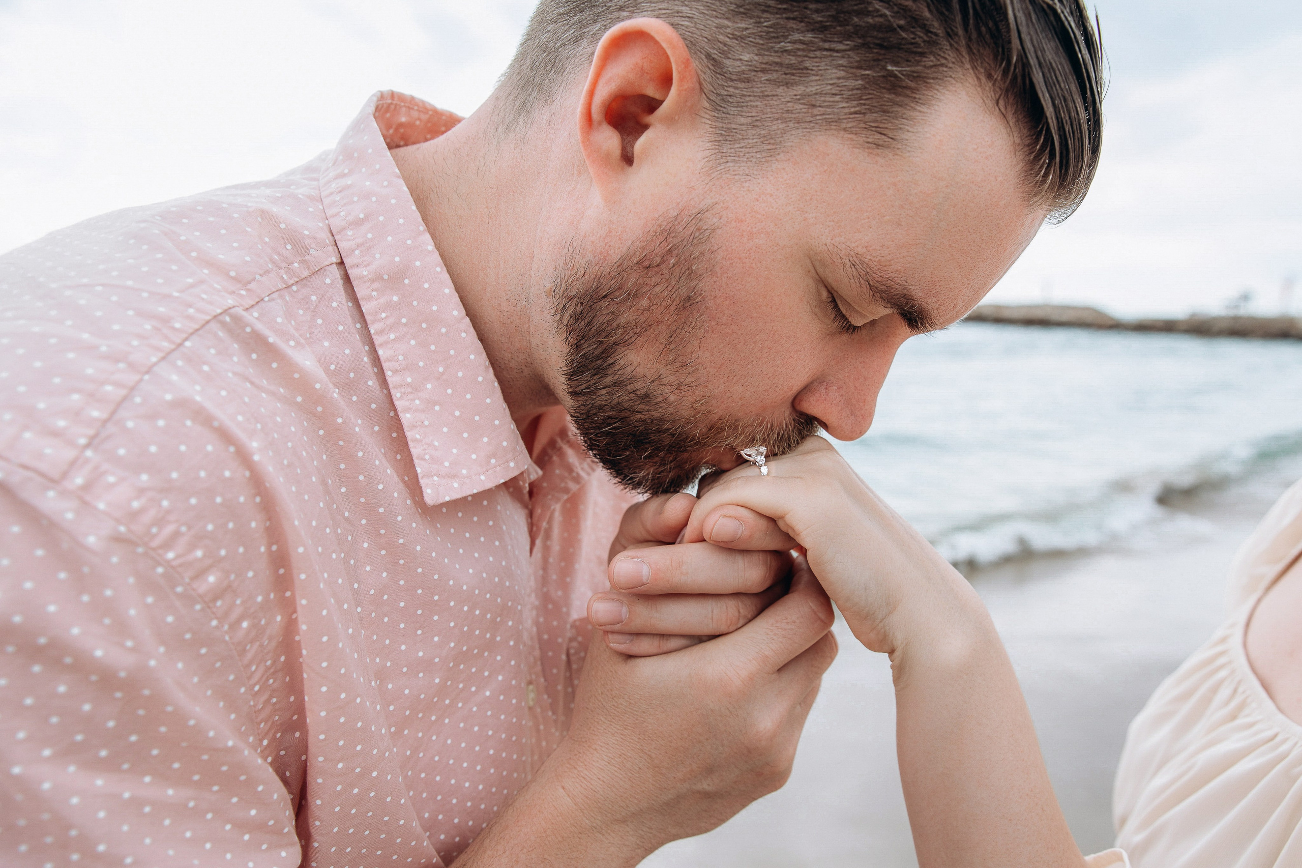 Close-up of a man kissing his partner’s hand by the sea during a proposal photoshoot.