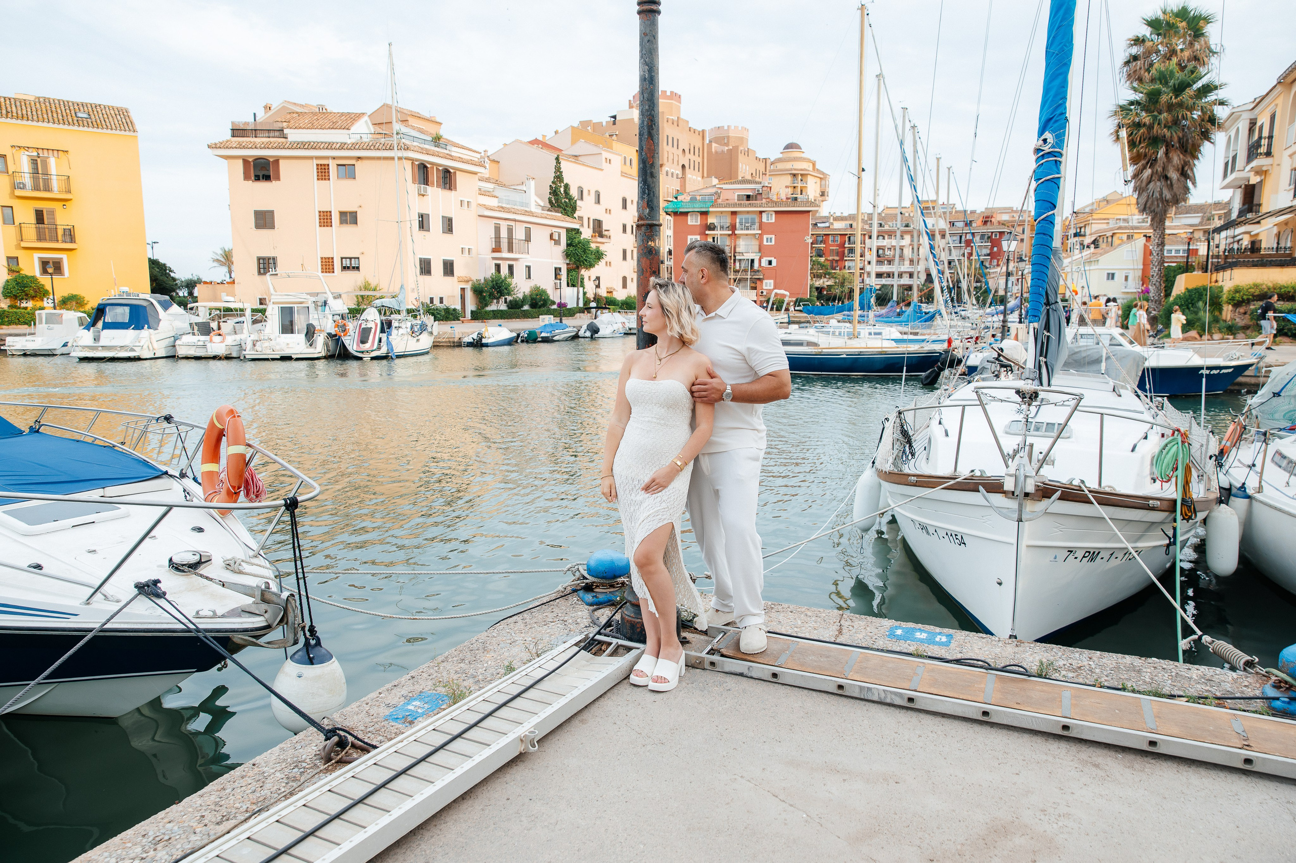 Couple embracing beside sailboats at Port Saplaya marina, with colourful waterfront buildings behind them.