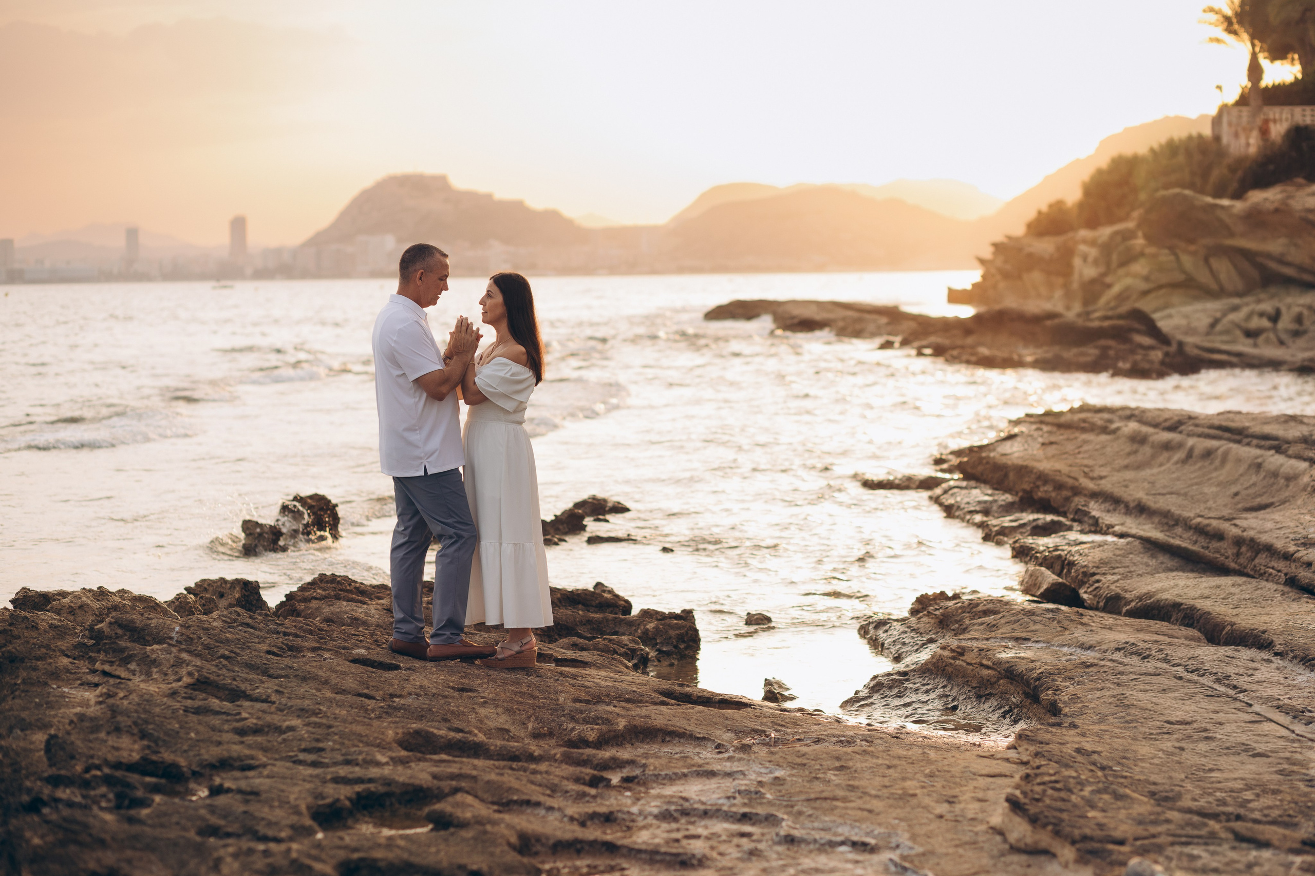 Couple embracing on a rocky shoreline in golden light during a beach proposal photoshoot.