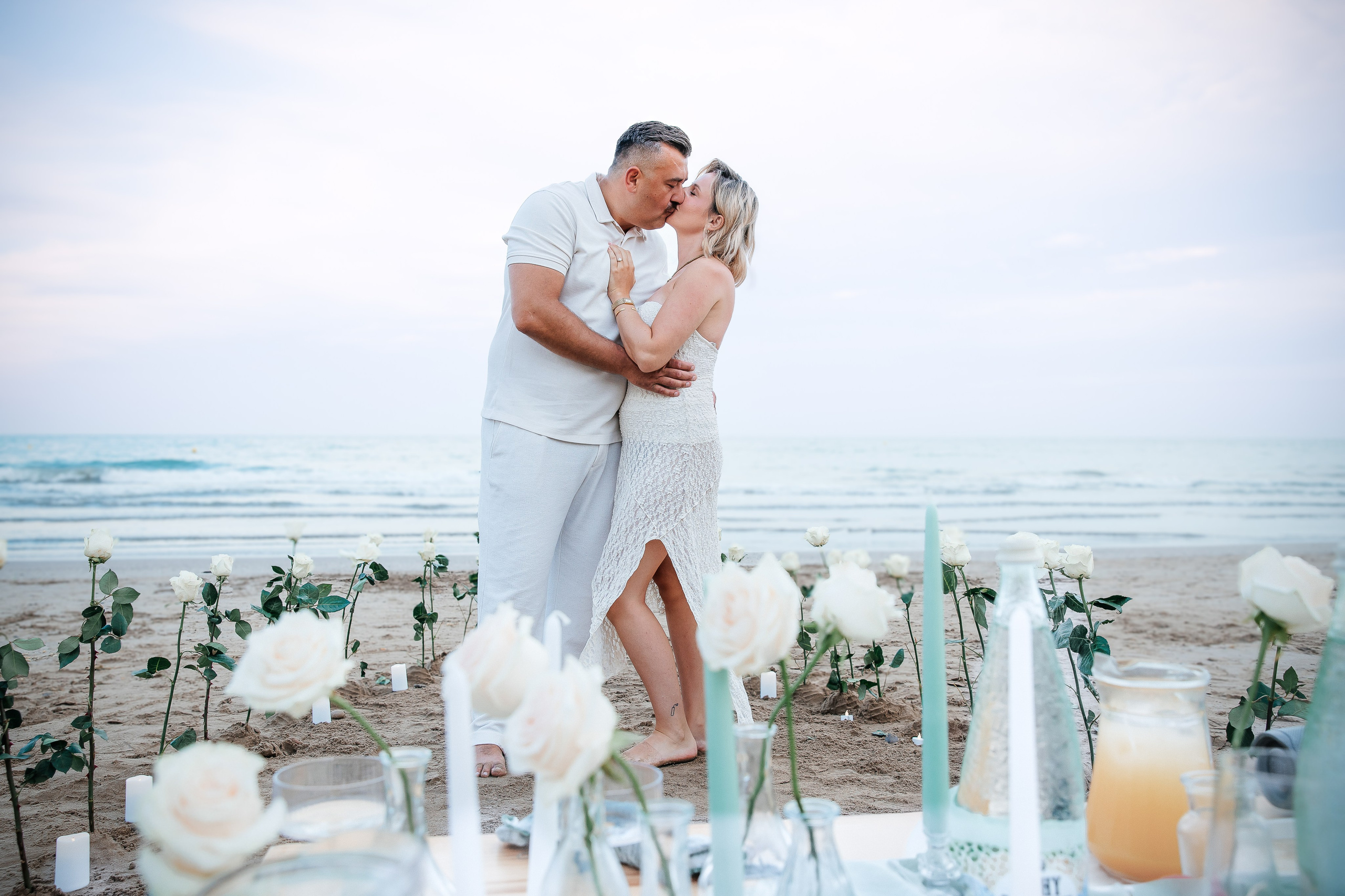 Couple kissing beside a seaside proposal setup decorated with white flowers and candles.