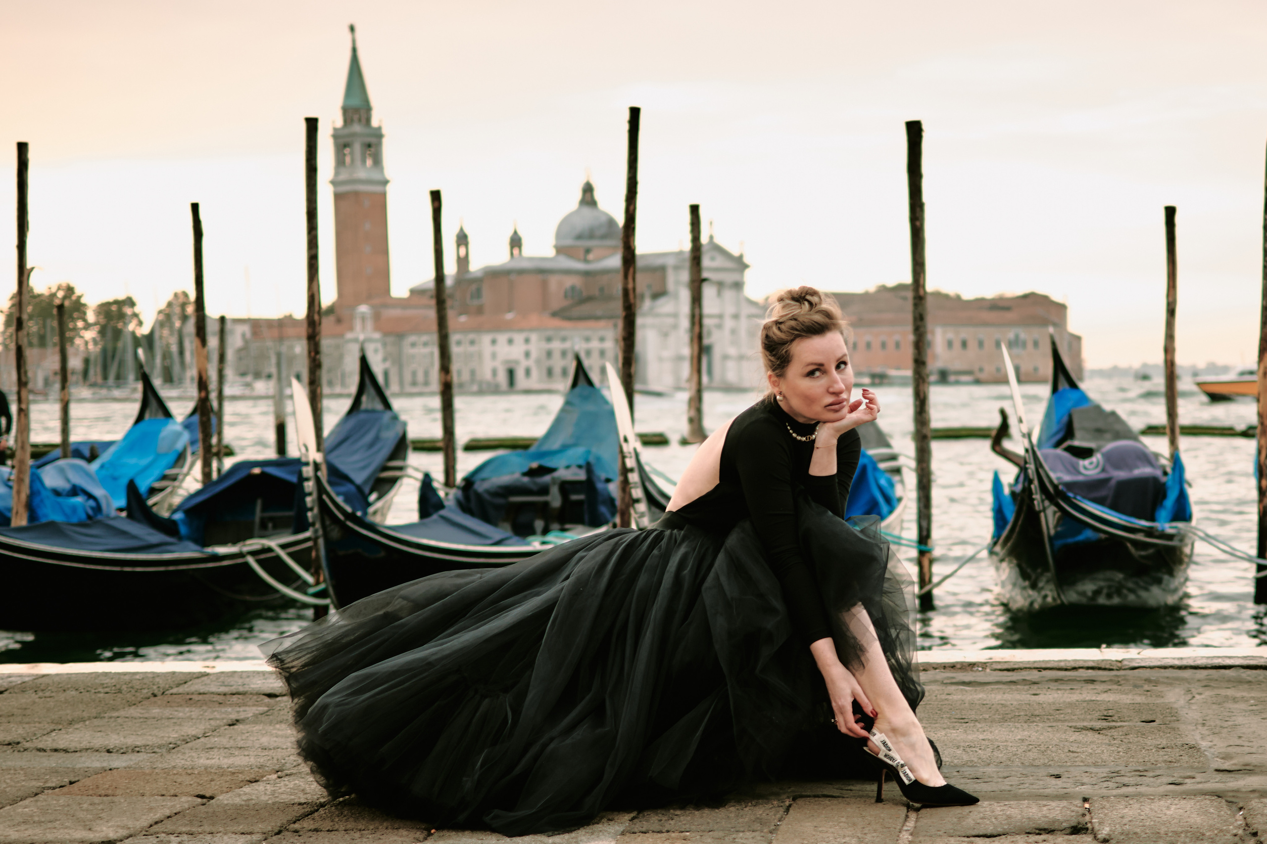 A young woman stands in the heart of San Marco Square, wearing a stunning black dress that accentuates her beauty. As she poses for the camera, the panoramic view of Laguna di Venezia provides a breathtaking backdrop. The intricate architecture of the surrounding buildings adds to the romantic and captivating atmosphere of the scene. The woman's hair blows gently in the warm breeze, and her smile radiates joy and happiness. This photo captures the essence of Venice's charm and timeless elegance. Fashion blogger shoot Venice. Portrait and Fashion Photographer in Venice, Italy. Instagram Photos with lifestyle photographer