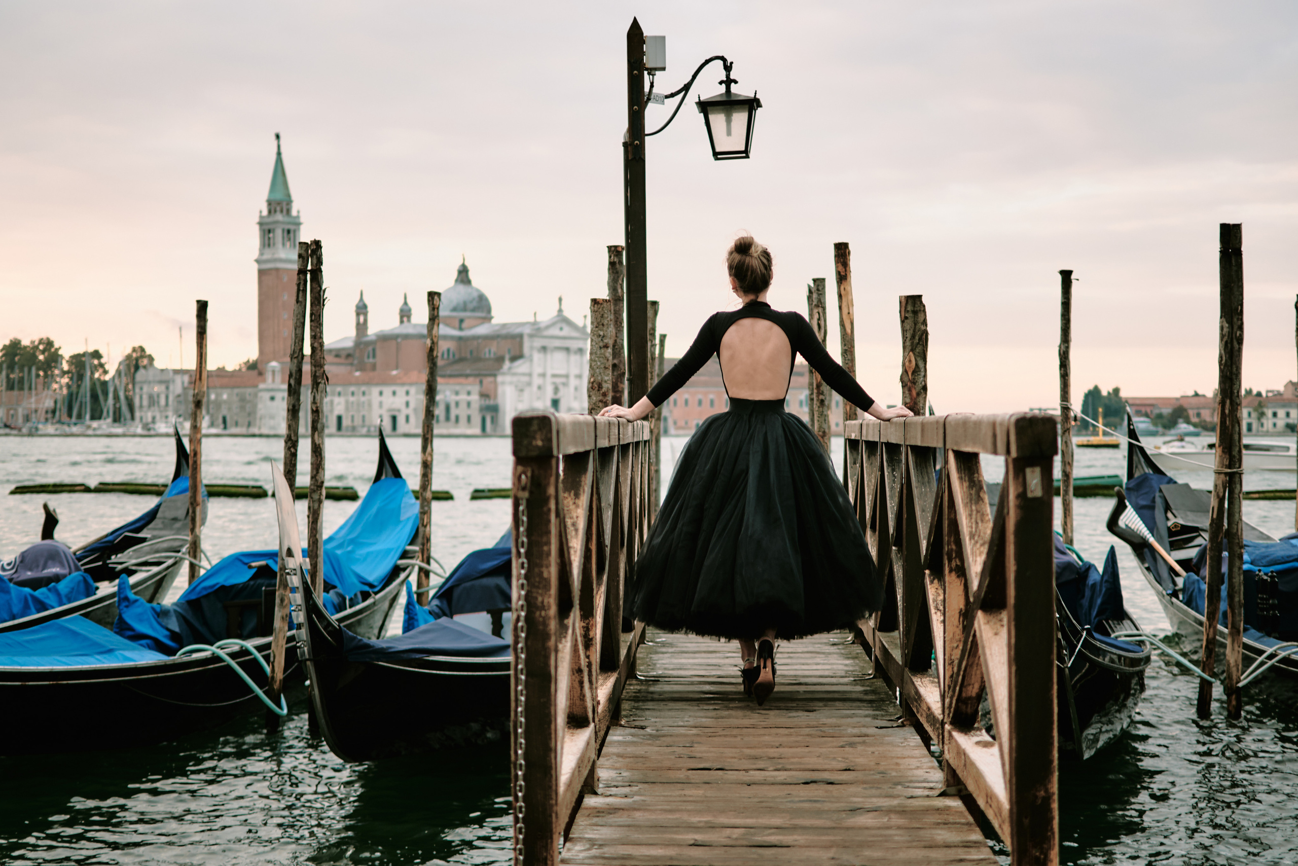 A woman stands in the heart of San Marco Square in Venice, wearing a stunning black dress that accentuates her beauty. The panoramic view of the Laguna in the background adds to the romantic atmosphere of the scene. The ornate architecture of the surrounding buildings is a testament to the city's rich history and culture. The woman's hair flows gently in the breeze as she gazes out at the stunning view, lost in thought. The vibrant energy of the bustling square is palpable, making for a truly memorable photo shoo