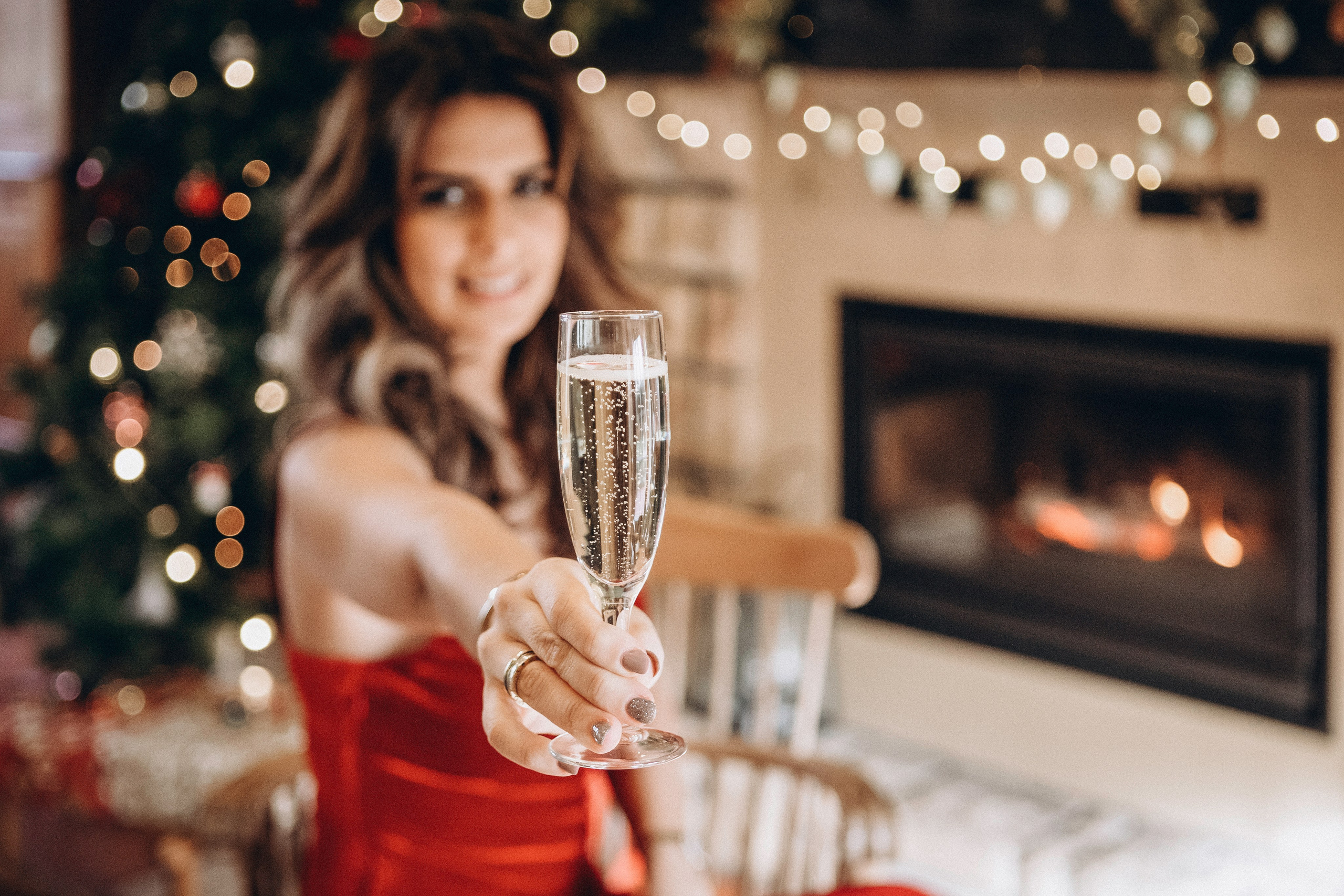 Femme tenant une coupe de champagne lors d’une séance photo de Noël chaleureuse en intérieur avec guirlandes et cheminée.