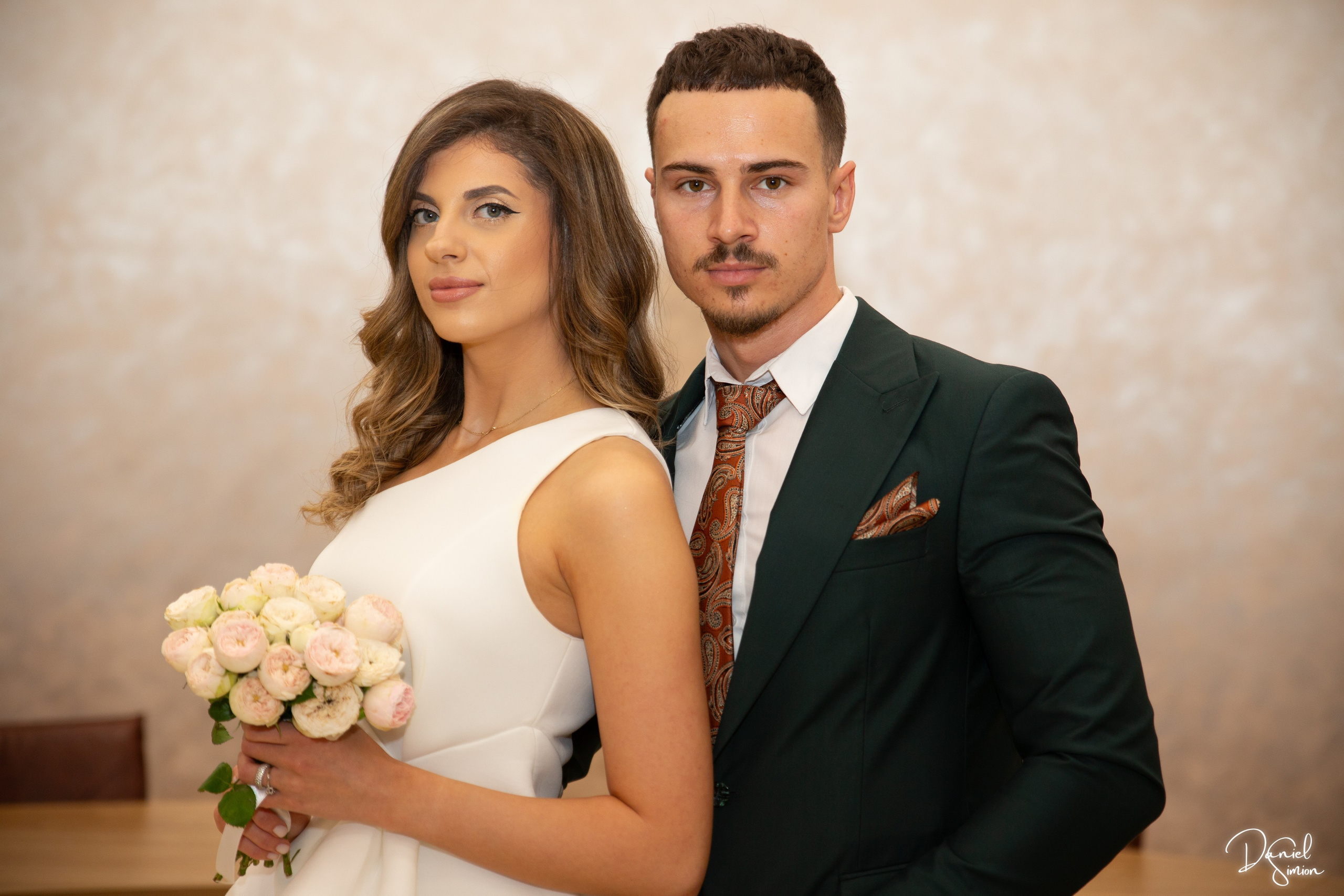 Bride in white dress holding pale rose bouquet and groom in dark suit with patterned tie standing indoors.