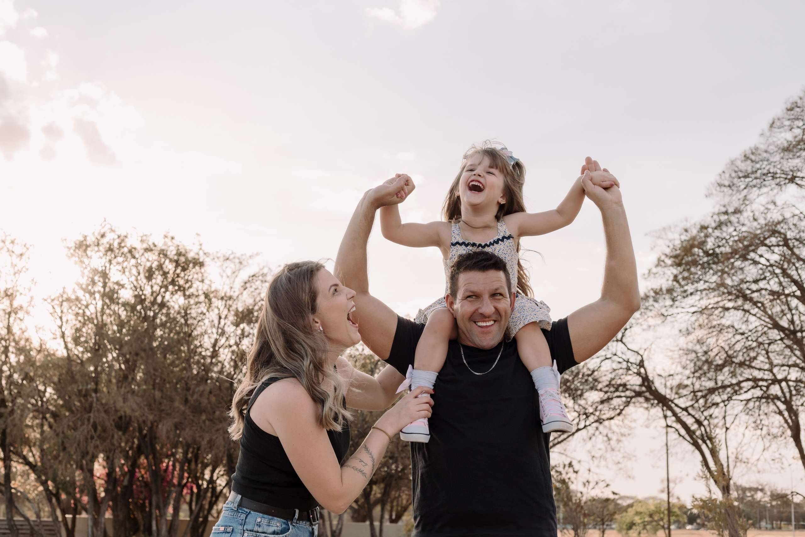 Fotografia de família com menina nos ombros do pai e olhar afetuoso da mãe