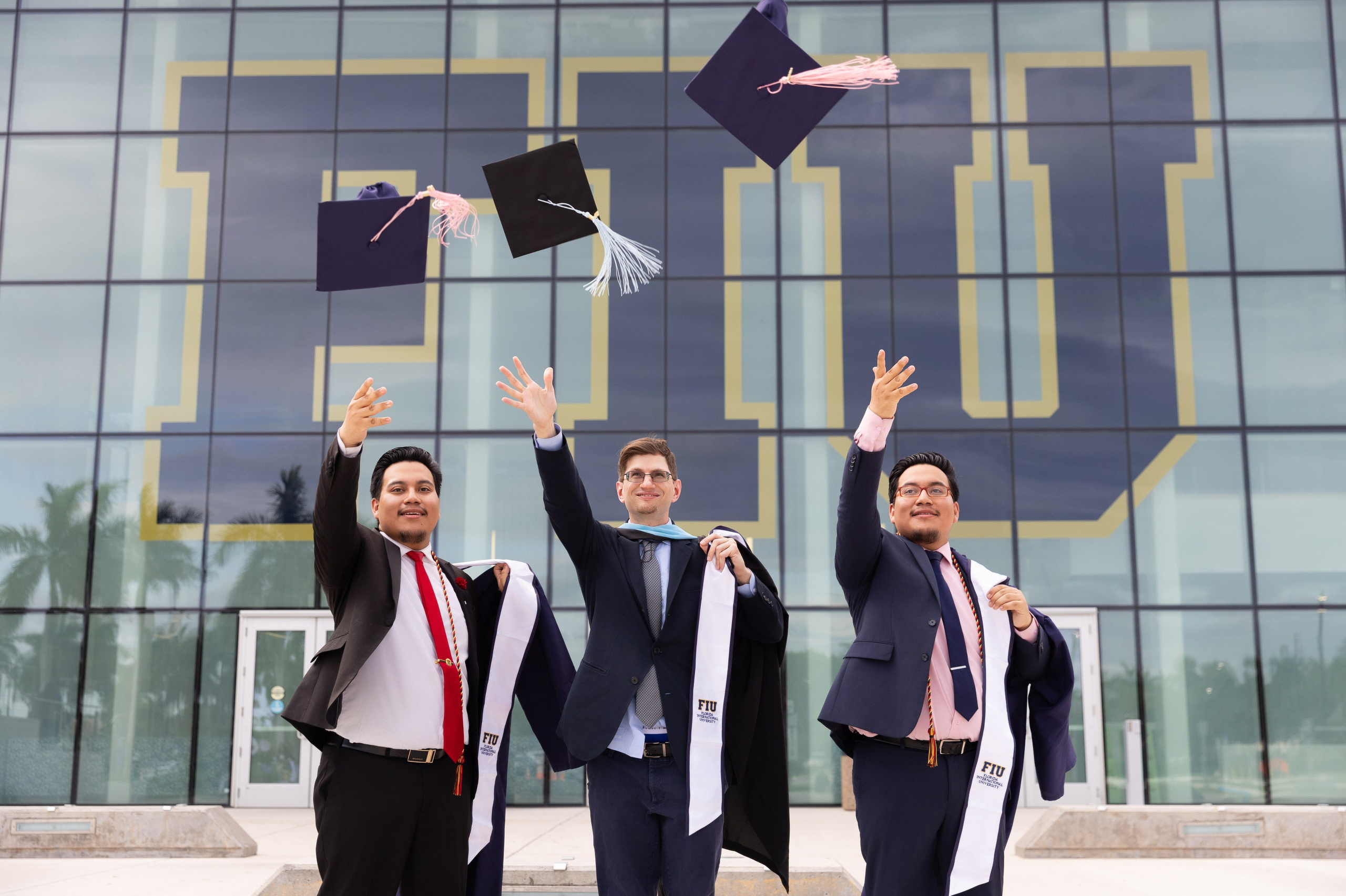 Three FIU music graduates toss their caps outside the campus building.