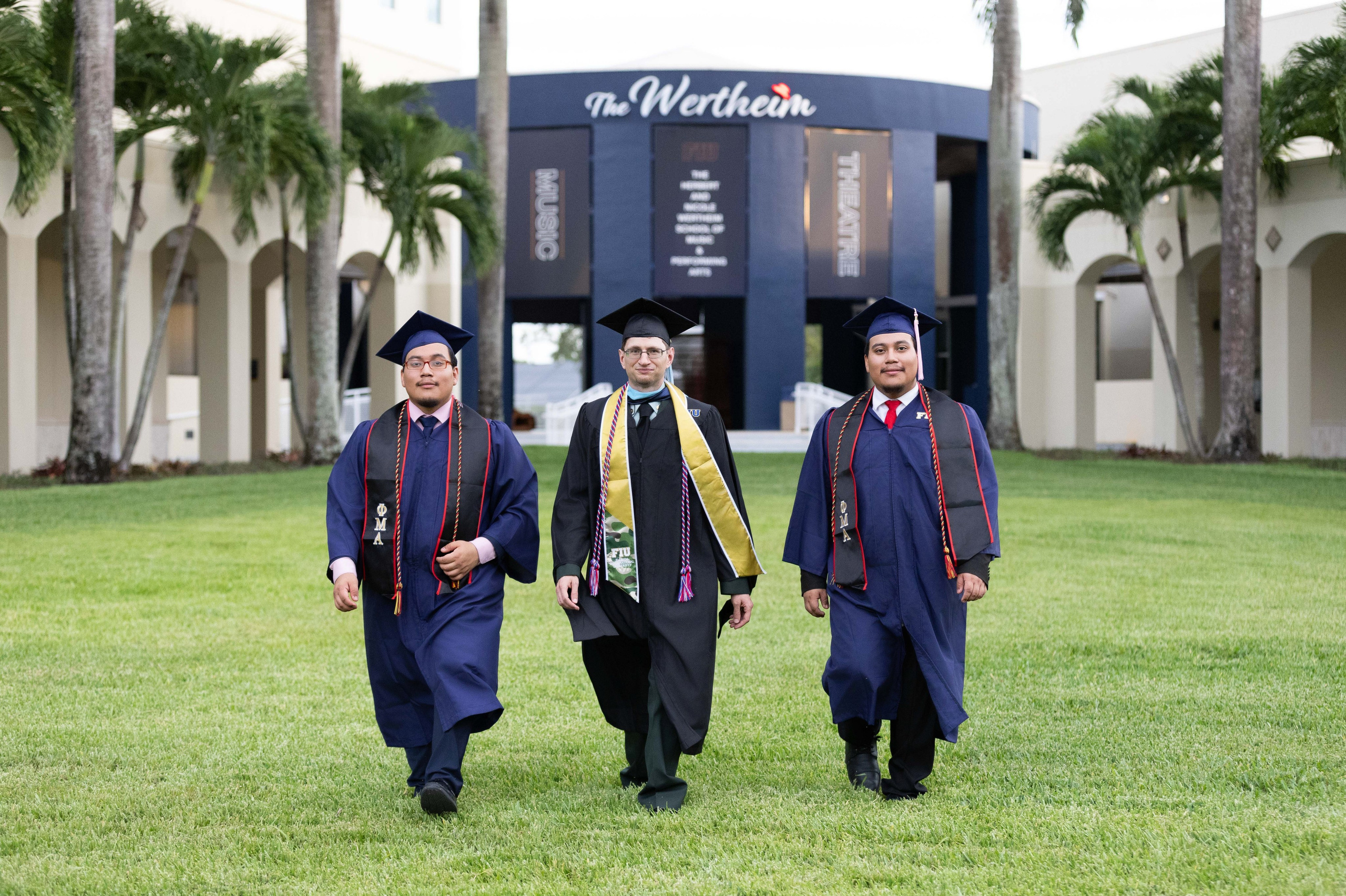 Three Wertheim School of Music graduates walk across the FIU campus lawn.
