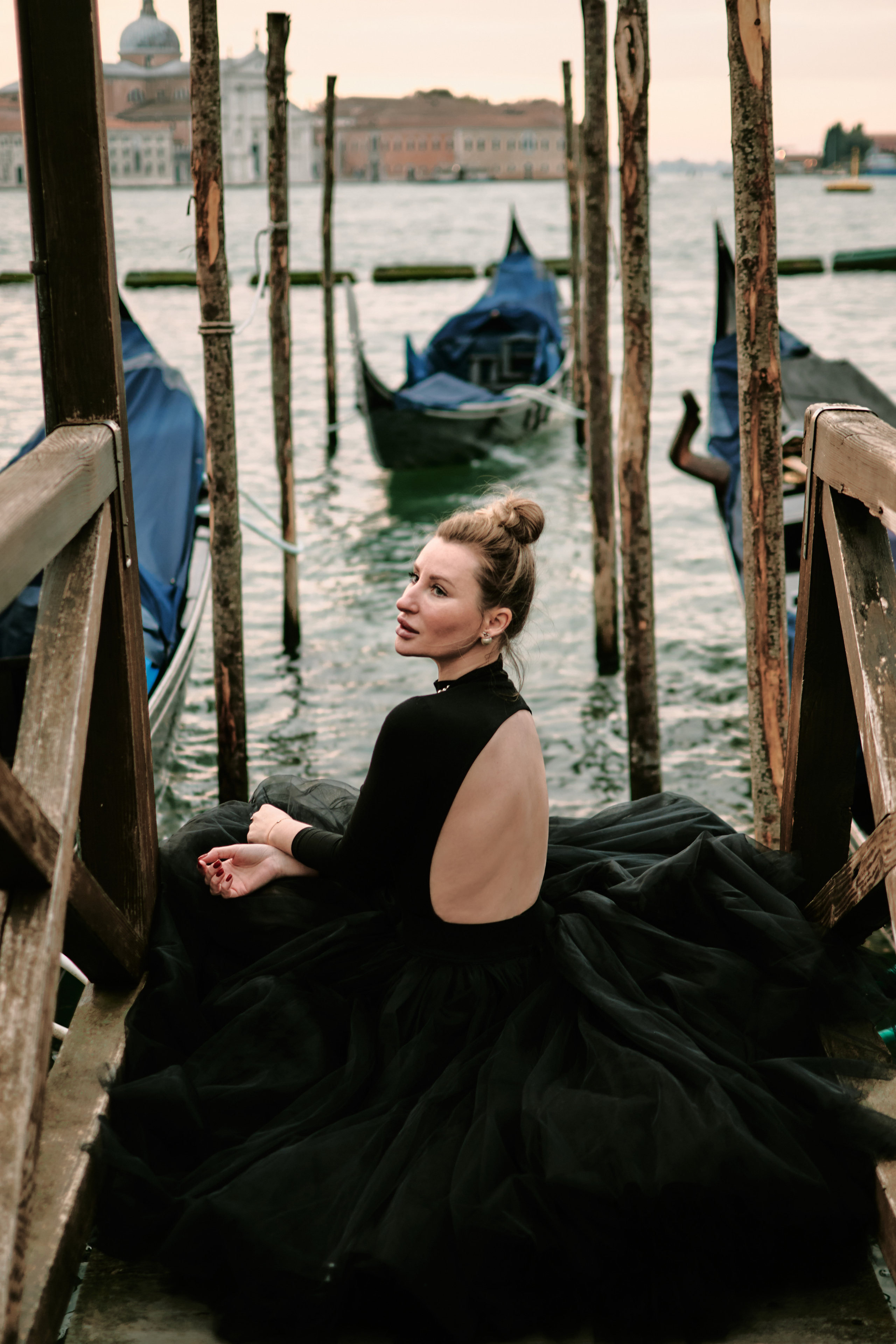 Portrait of a young woman in a black dress standing on San Marco Square, Venice, with a breathtaking panoramic view of the Laguna in the background. The woman has long brown hair styled in loose waves, and she is looking dreamily towards the view. The square is bustling with people, and the stunning architecture of the surrounding buildings adds to the romantic atmosphere of the location