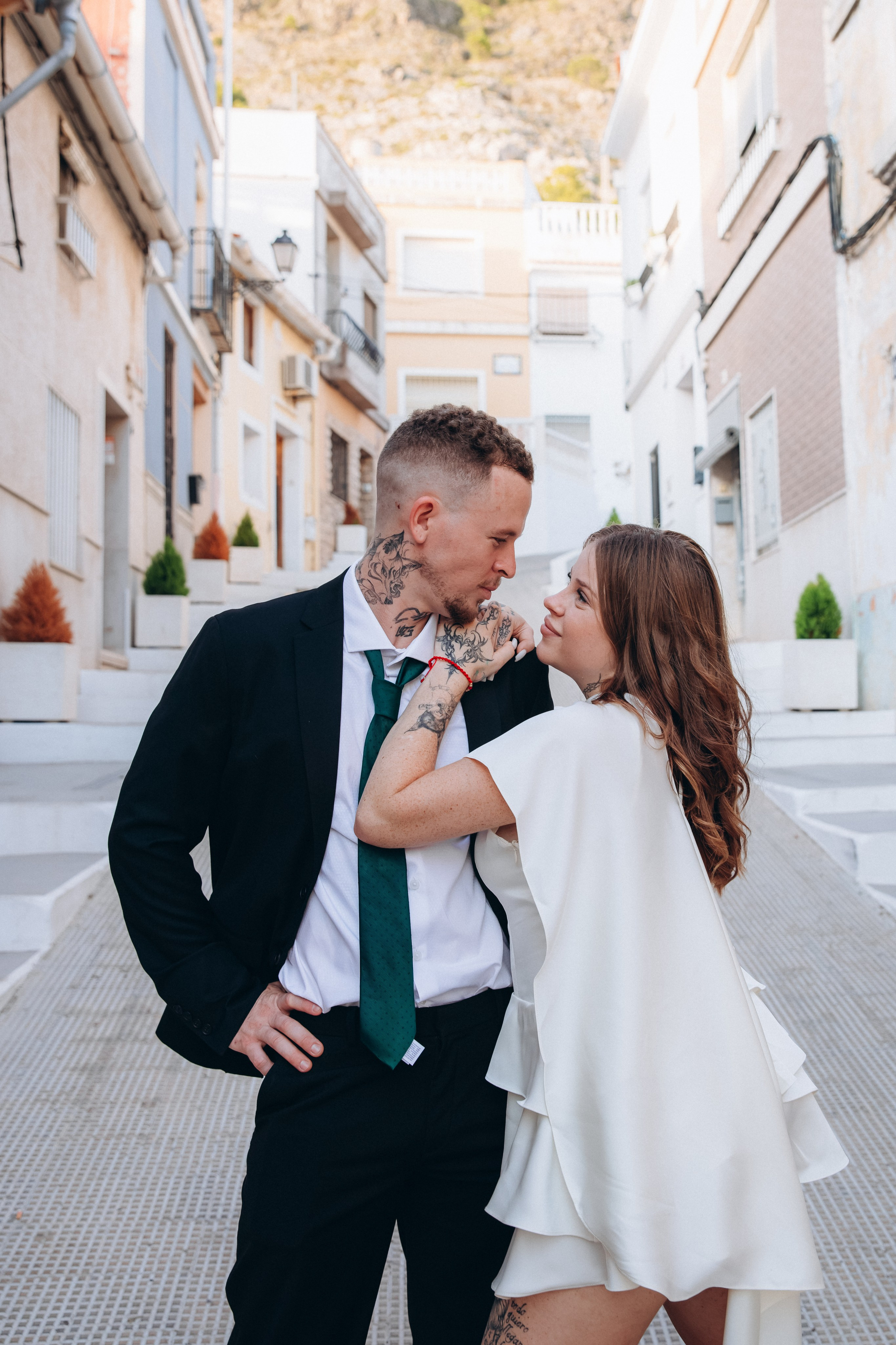 Couple sharing a kiss on a narrow city street, wearing a dark suit and a short white dress.