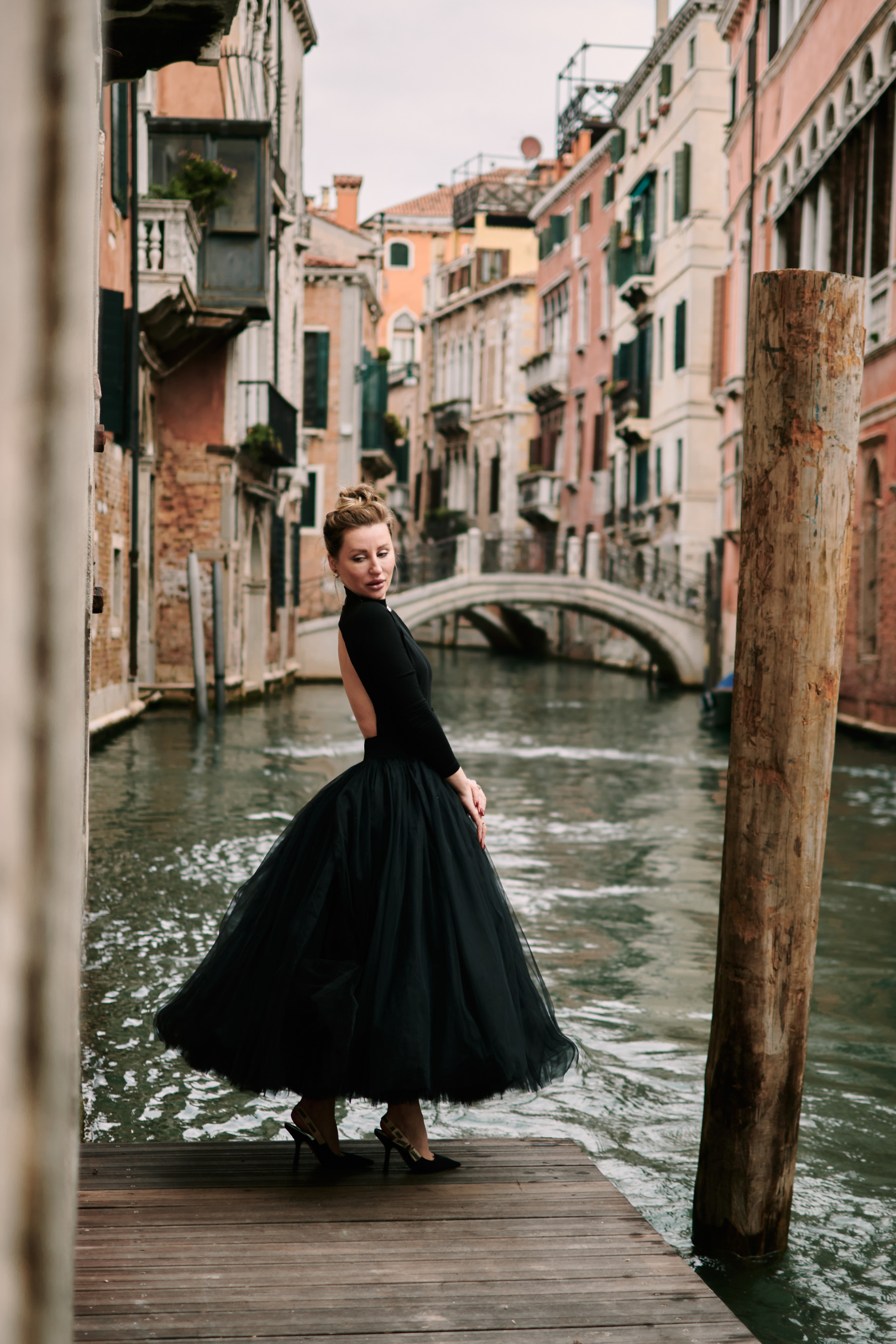A captivating young woman in a flowing black dress stands on a narrow bridge, framed by the intricate stonework of the arches above her. The scene is full of mystery and romance, with the gentle canal waters below and the timeless beauty of Venice's architecture surrounding her