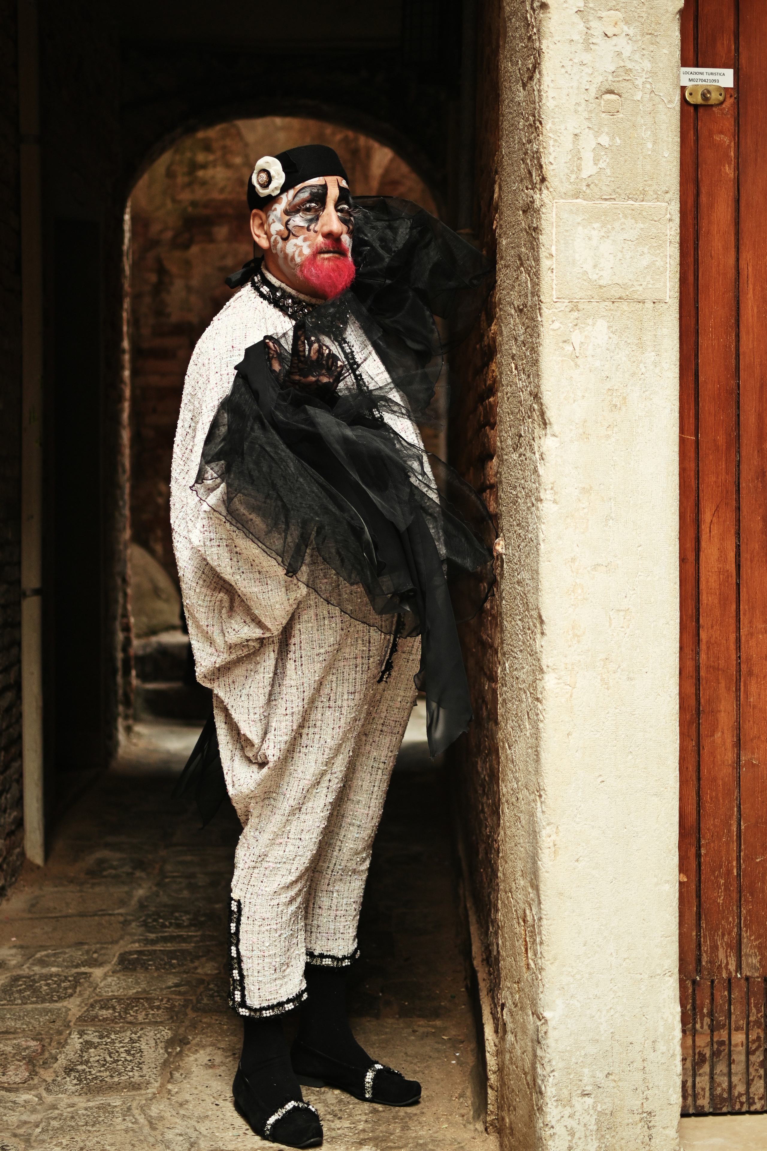 People wearing colorful costumes and masks at Venice Carnival. Colorful costumes and masks of the Carnival in Venice, Italy, with people walking on a crowded street. A man in a white hat and black cloak stands in the foreground holding a red and black mask. Buildings with ornate architecture and colorful flags are visible in the background.