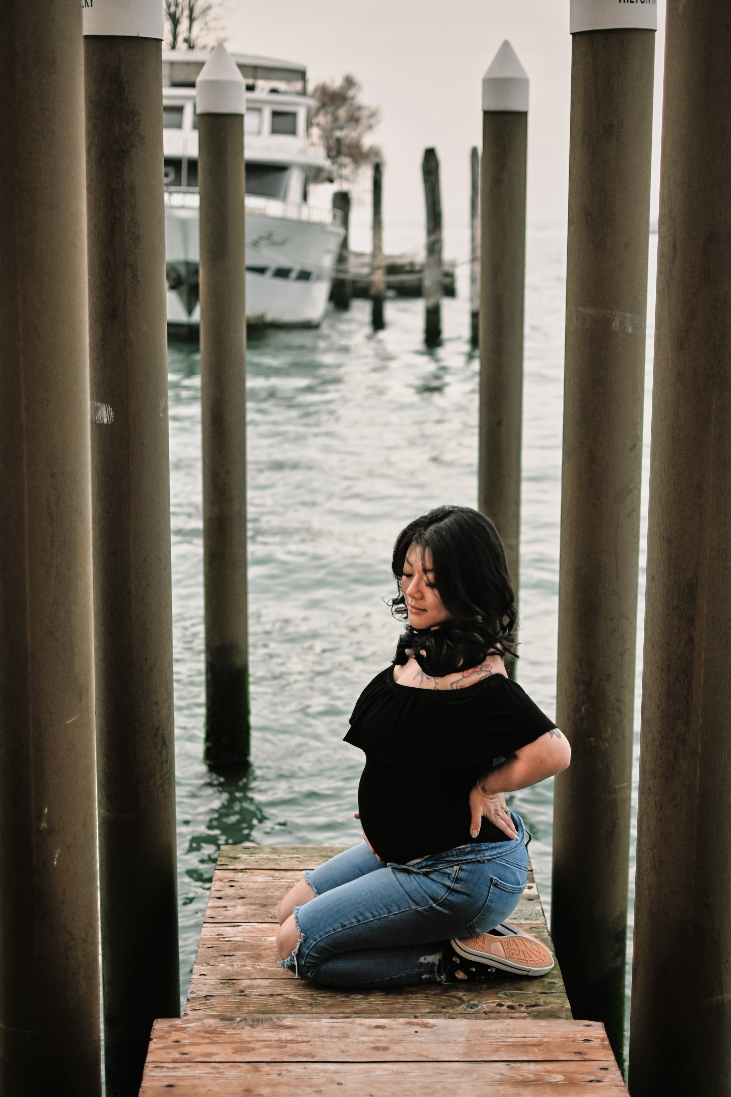 A pregnant young woman stands on a bridge in Venice, wearing a pair of comfortable jeans and a flowing black blouse. Her hair is pulled back in a loose bun, and she gazes out at the bustling canal below. The sunlight glimmers on the water, casting a warm glow over the scene. In the background, ancient buildings and colorful boats lend a sense of history and romance to the setting. The woman's pose is relaxed yet confident, conveying her excitement and anticipation for the new life growing inside her.
