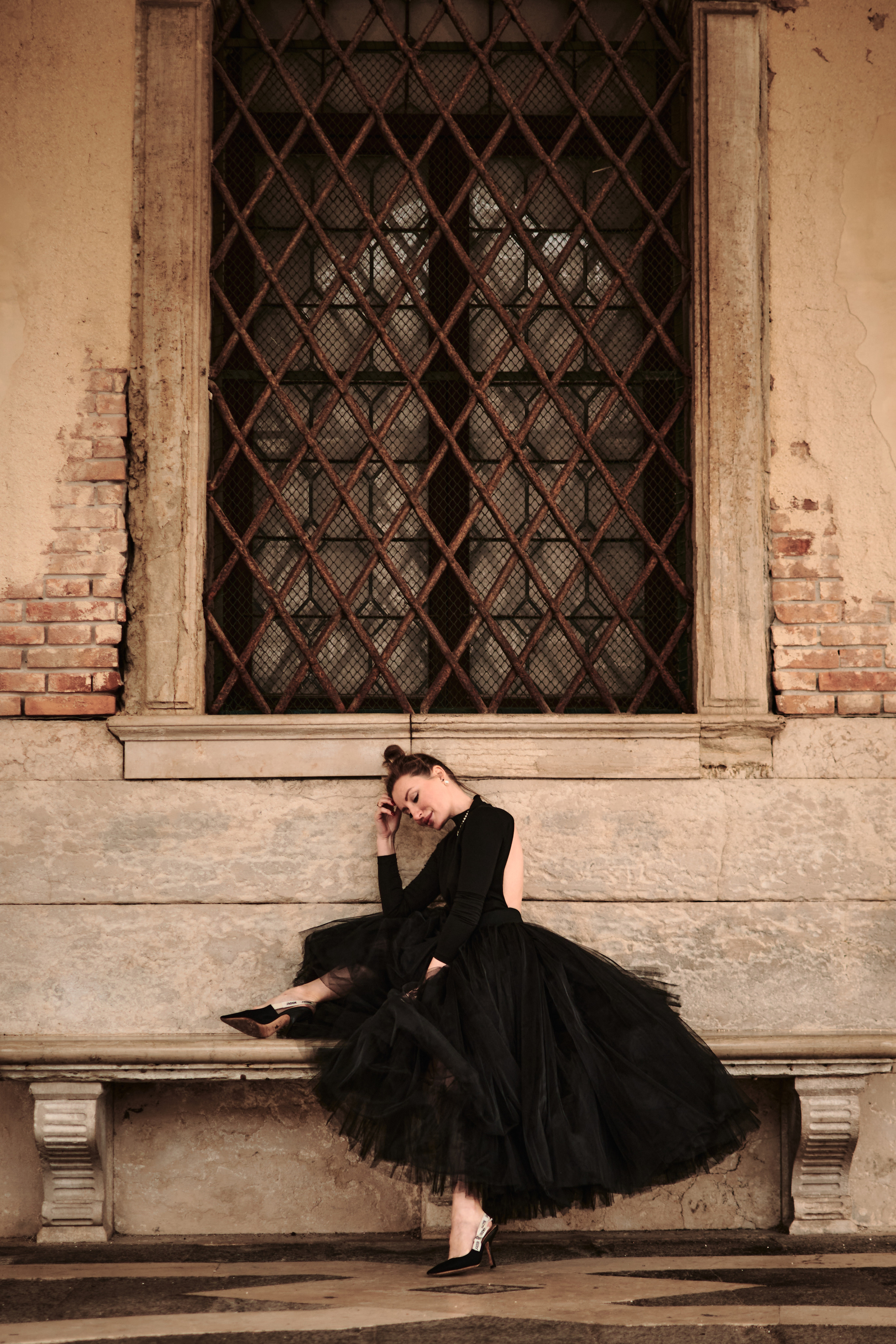 This portrait captures the timeless elegance and allure of Venice, with a young woman in a flowing black dress standing on a narrow bridge overlooking the canal below. The intricate stonework of the bridge's arches creates a unique frame within the photo, drawing the viewer's eye to the woman's captivating presence