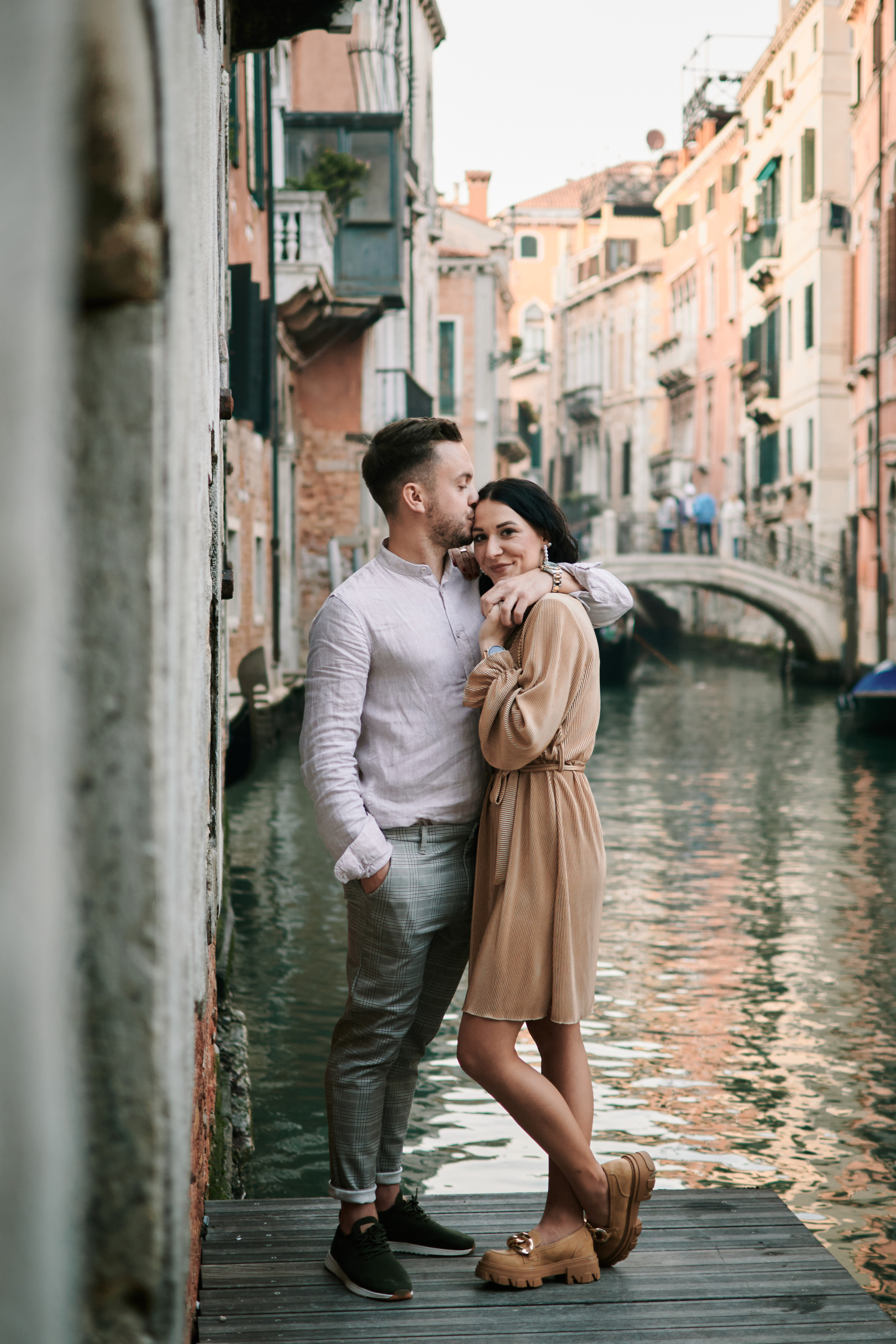 A couple stands hand in hand on a narrow cobblestone street in Venice. The winding alleyway is illuminated by the warm glow of street lamps and the occasional light from a nearby shop window. The couple is dressed in stylish attire, the man in a sharp suit and the woman in a flowing gown. They gaze lovingly into each other's eyes, lost in their own world amidst the charm and romance of the ancient Italian city. In the background, the muted sounds of Venetian life can be heard, adding to the atmosphere of peacefulness and intimacy.