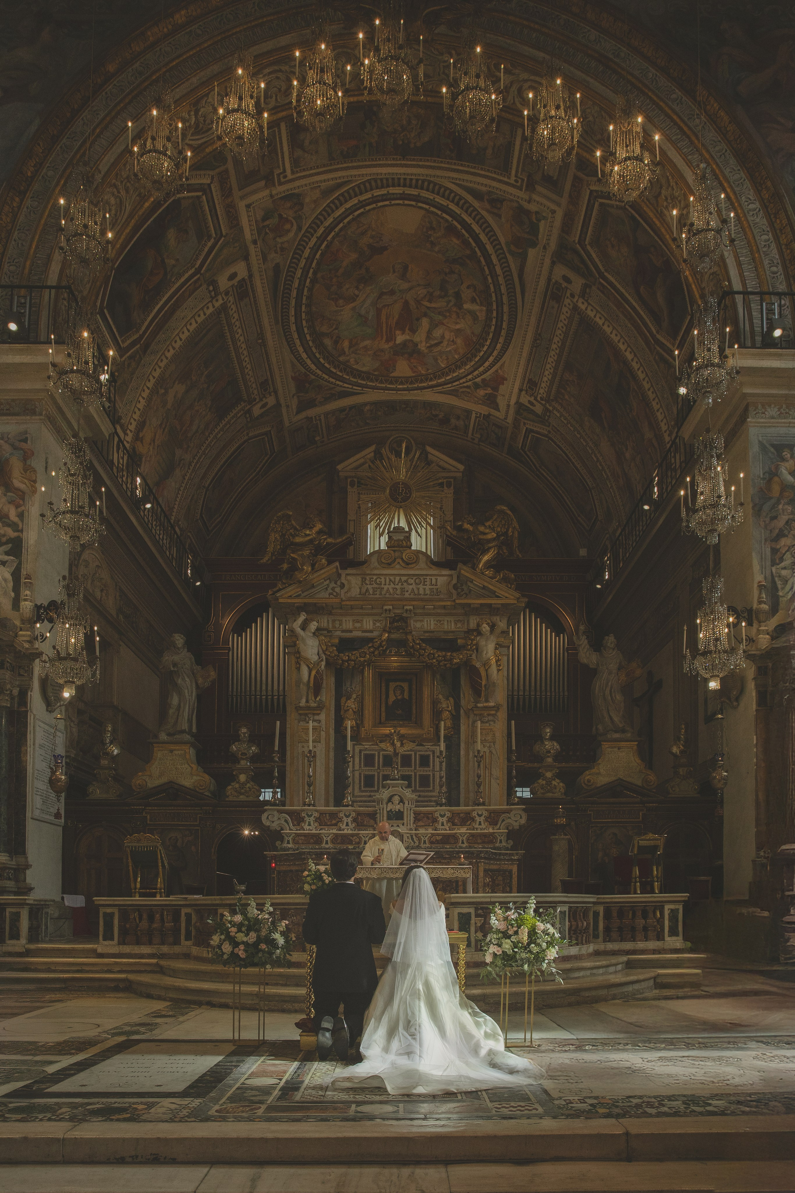 Bride and groom kneeling at the altar in a magnificent baroque church in Rome during an intimate Catholic wedding ceremony, with floral decorations and ornate frescoes.