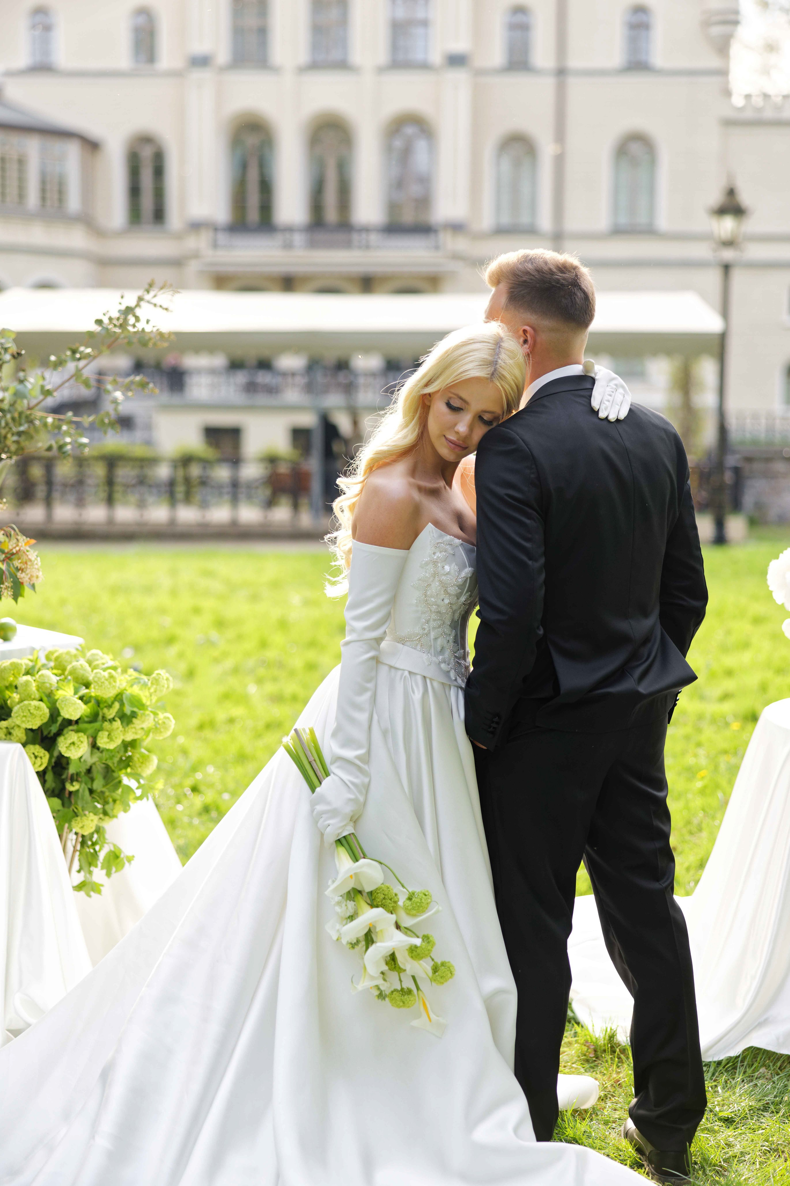 "Bride in a white gown holding a bouquet, leaning her head on the groom's shoulder in front of a villa."