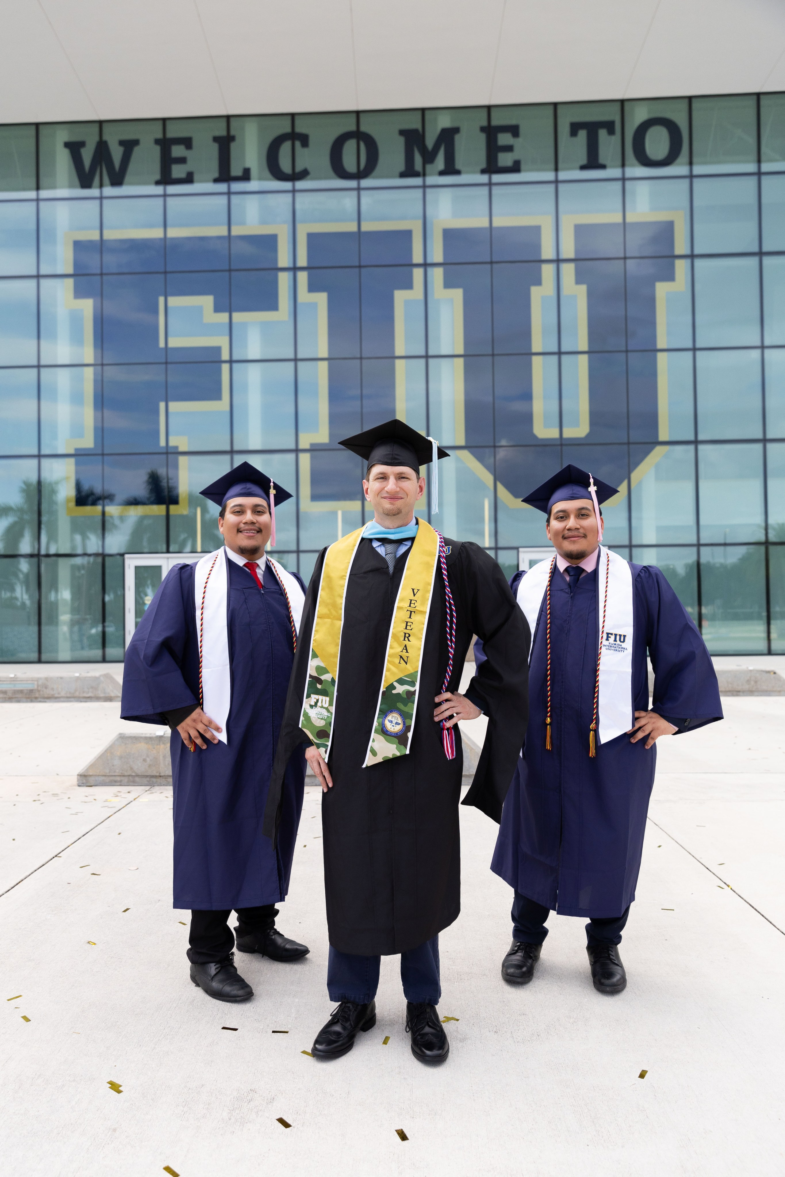 Darren and two classmates wear graduation gowns outside the FIU building.