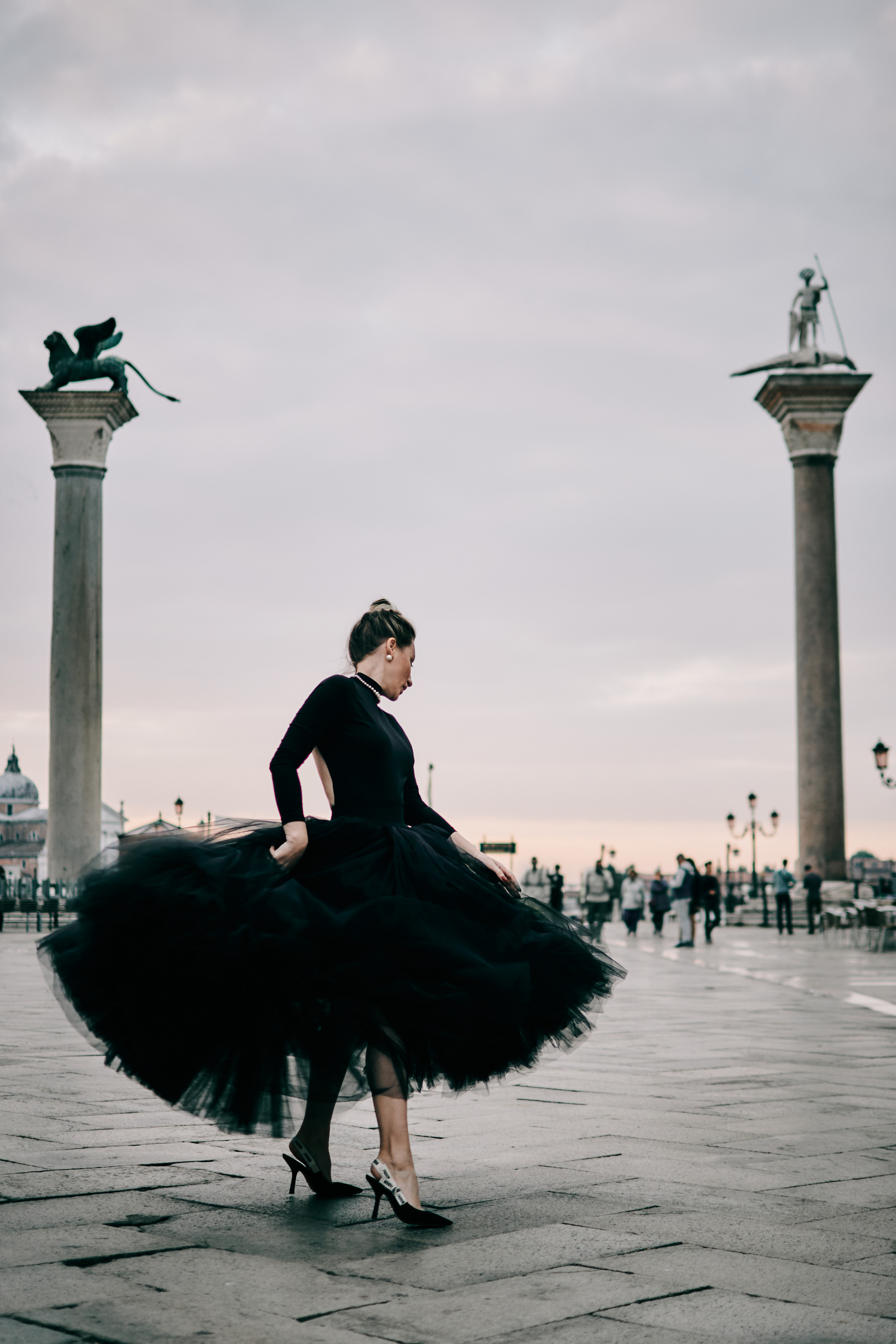 A young woman stands in front of the Columns of San Marco and San Todaro on San Marco Square in Venice. The sun shines brightly, casting a warm glow on the magnificent architecture and illuminating the intricate details of the columns. The woman wears a flowing black dress that billows gently in the breeze, adding to the romantic and ethereal atmosphere of the photo. She gazes off into the distance, lost in thought or perhaps captivated by the beauty of her surroundings. Fashion blogger shoot Venice. Portrait and Fashion Photographer in Venice, Italy. Instagram Photos with lifestyle photographer