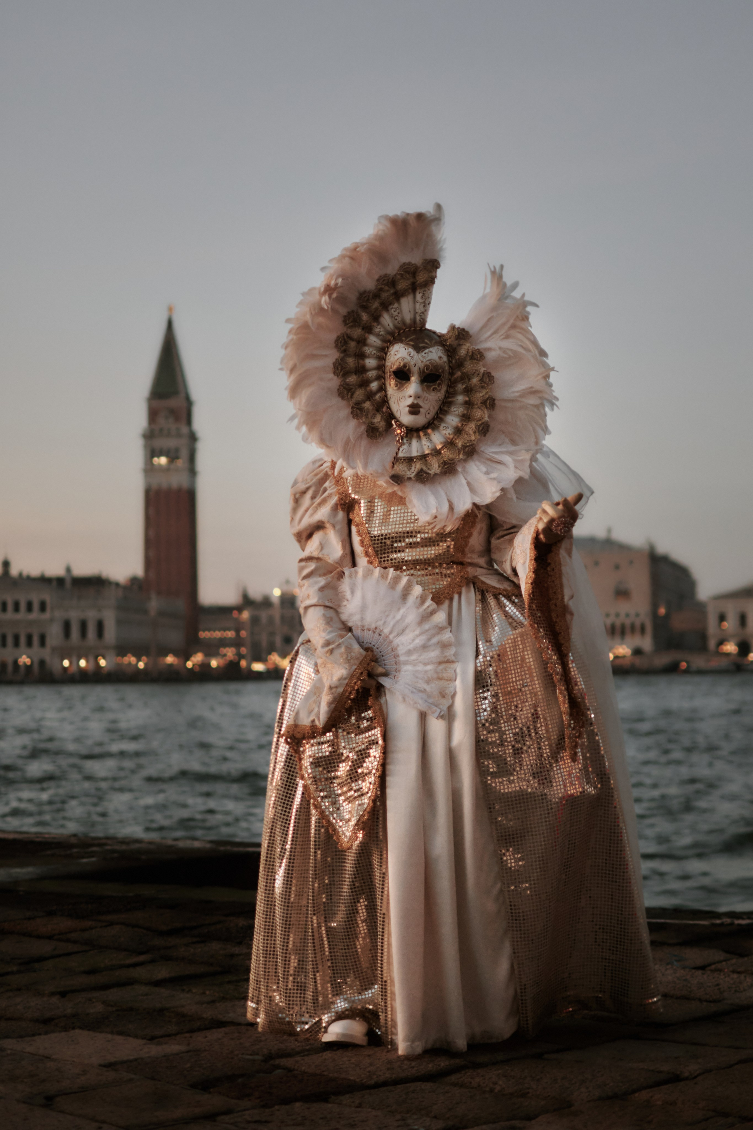 A couple wearing matching costumes and masks at Venice Carnival. Against the stunning backdrop of San Marco Square, a couple stands hand in hand, their love for each other evident in their smiles and the way they look at each other. The Columns of San Marco and San Todaro frame the couple, adding to the drama and beauty of the photo.