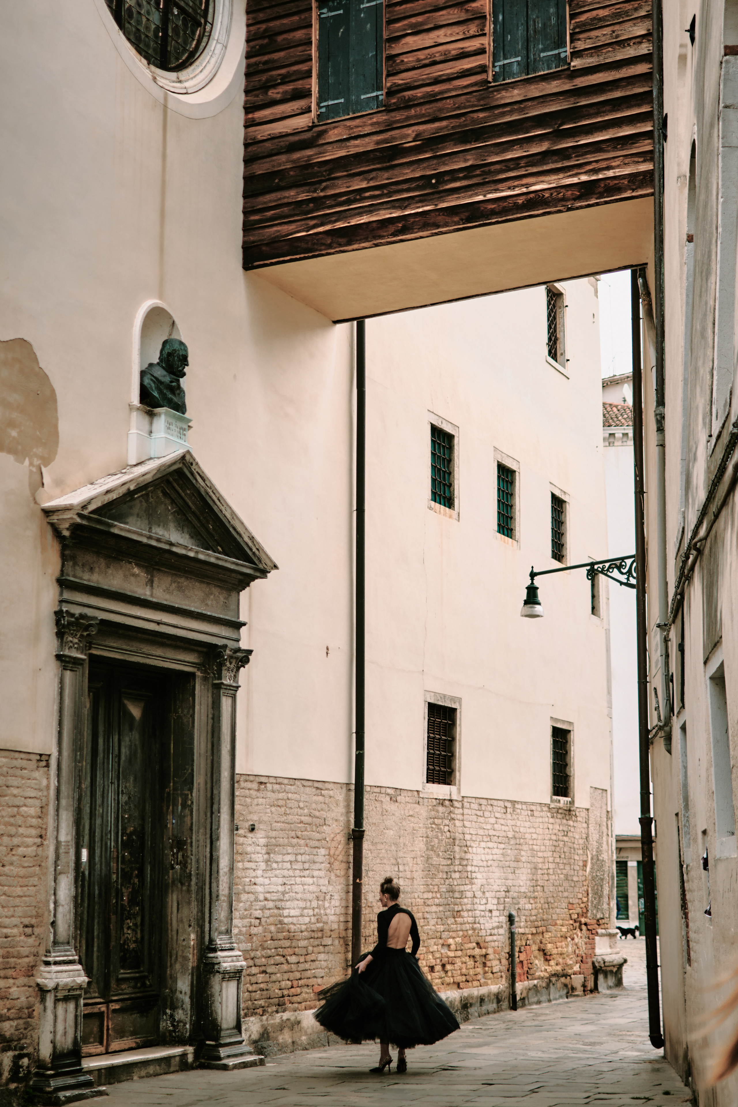 This portrait captures the timeless elegance and allure of Venice, with a young woman in a flowing black dress standing on a narrow bridge overlooking the canal below. The intricate stonework of the bridge's arches creates a unique frame within the photo, drawing the viewer's eye to the woman's captivating presence.