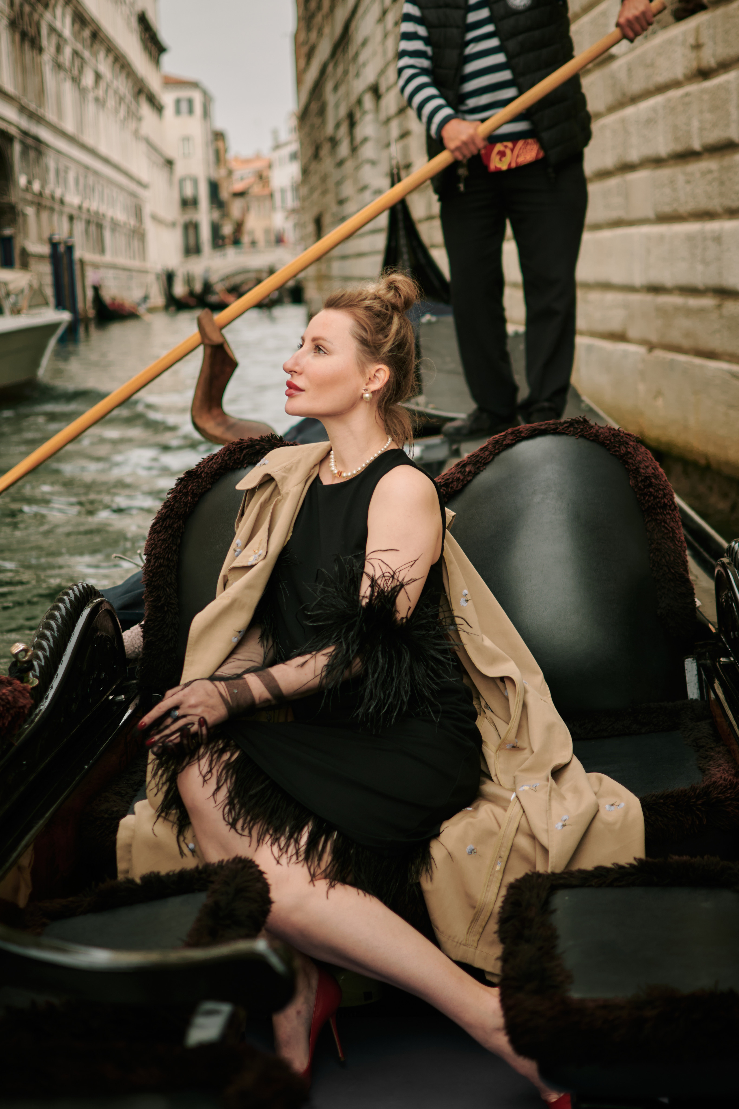 A young woman in a black dress sits on a gondola as it glides through the narrow streets of Venice. She gazes off into the distance, lost in thought, as the sunlight dances on the water around her. The picturesque buildings of the city rise up on either side, their pastel colors and ornate details adding to the romantic atmosphere. The woman's elegant dress and captivating presence seem perfectly at home in this enchanting setting, creating a sense of mystery and allure that draws the viewer. Fashion blogger shoot Venice. Portrait and Fashion Photographer in Venice, Italy. Instagram Photos with lifestyle photographer