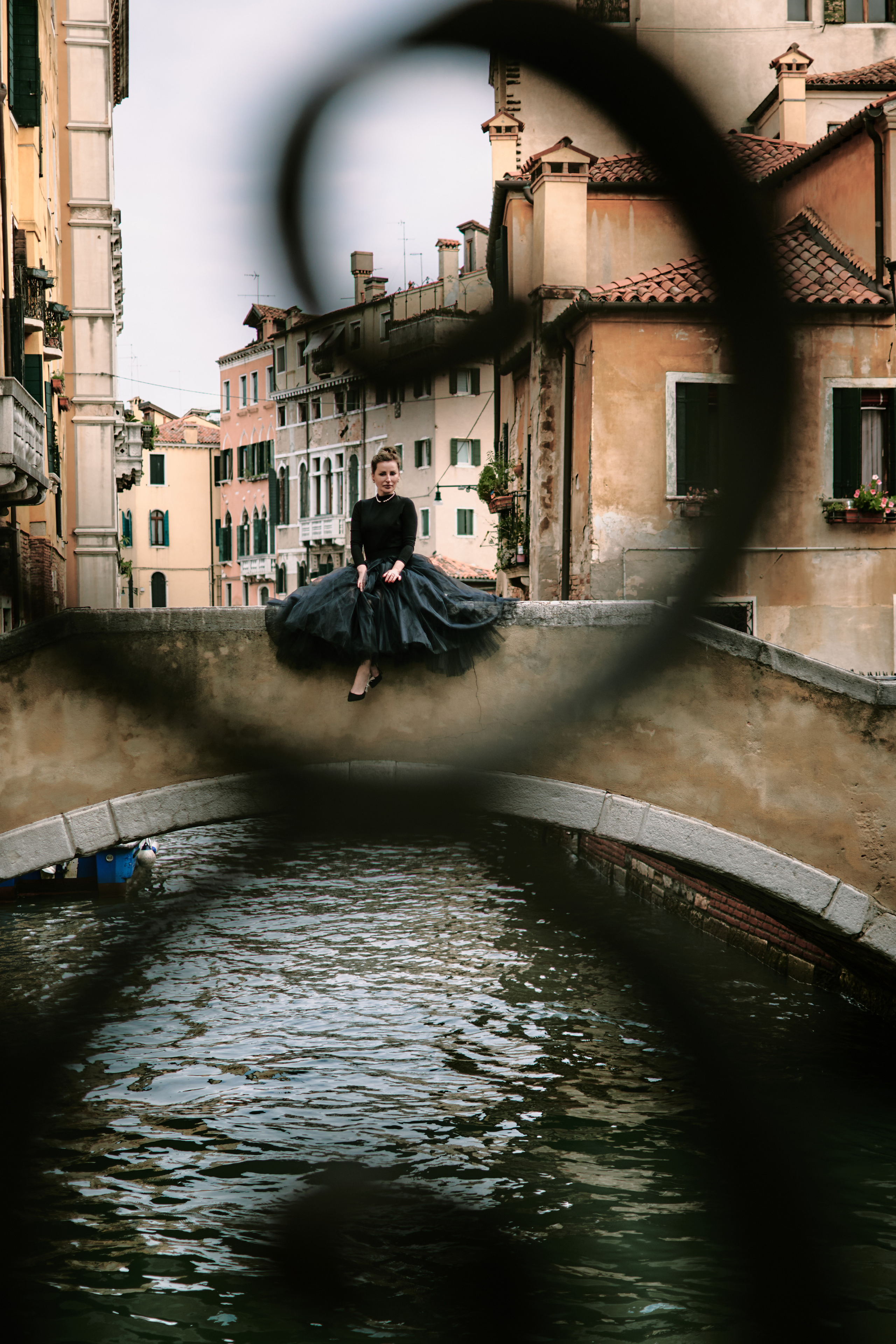 A captivating young woman in a flowing black dress stands on a narrow bridge, framed by the intricate stonework of the arches above her. The scene is full of mystery and romance, with the gentle canal waters below and the timeless beauty of Venice's architecture surrounding her