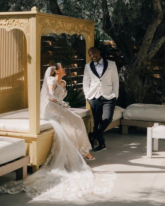 A group wedding photo with the bride and groom sharing a kiss in the foreground, surrounded by smiling guests, with tall palm trees under a bright sky.
